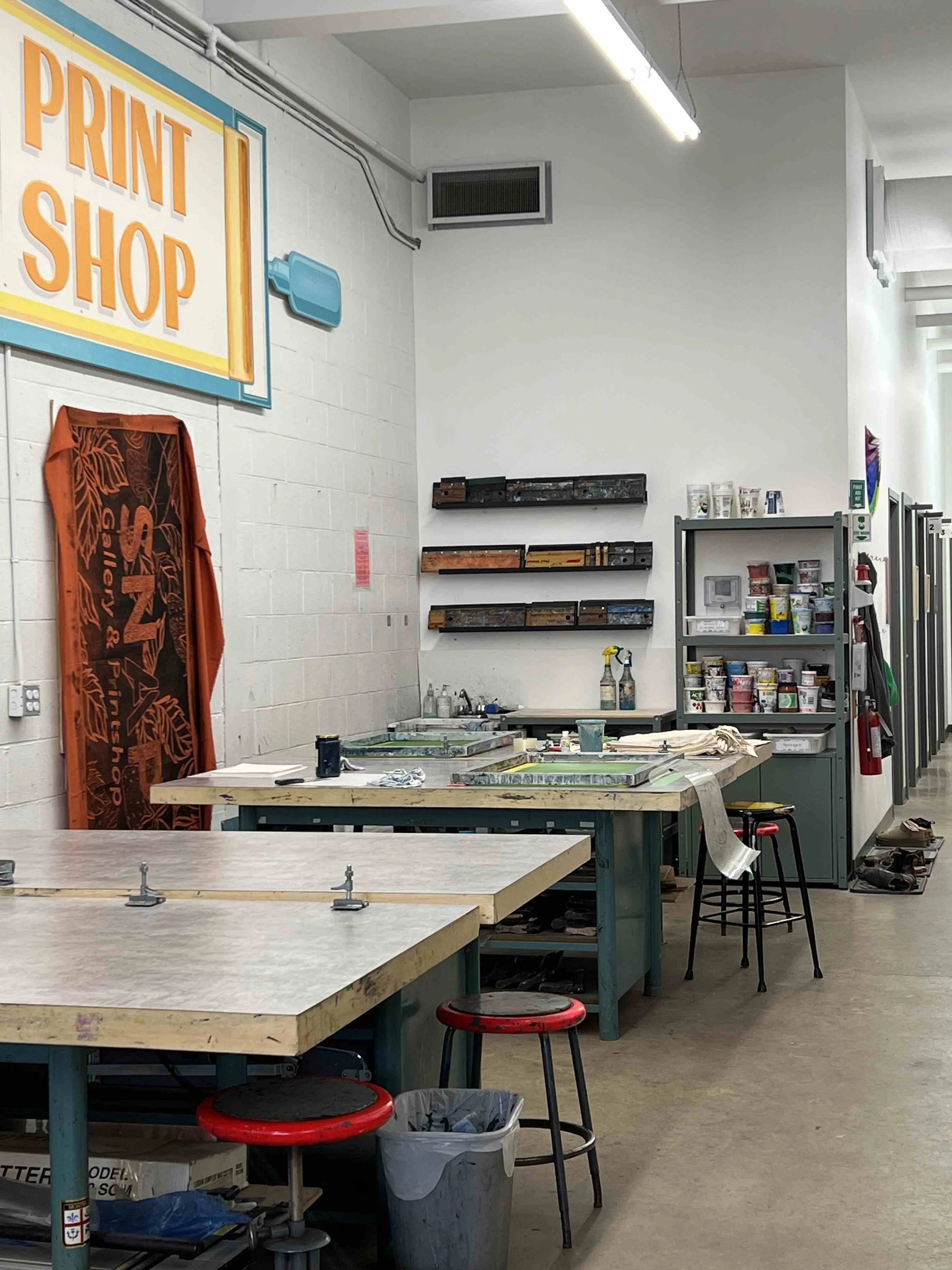 several screen printing stations sit under a sign reading "print shop" with squeegees and paint pots near a sink in the background