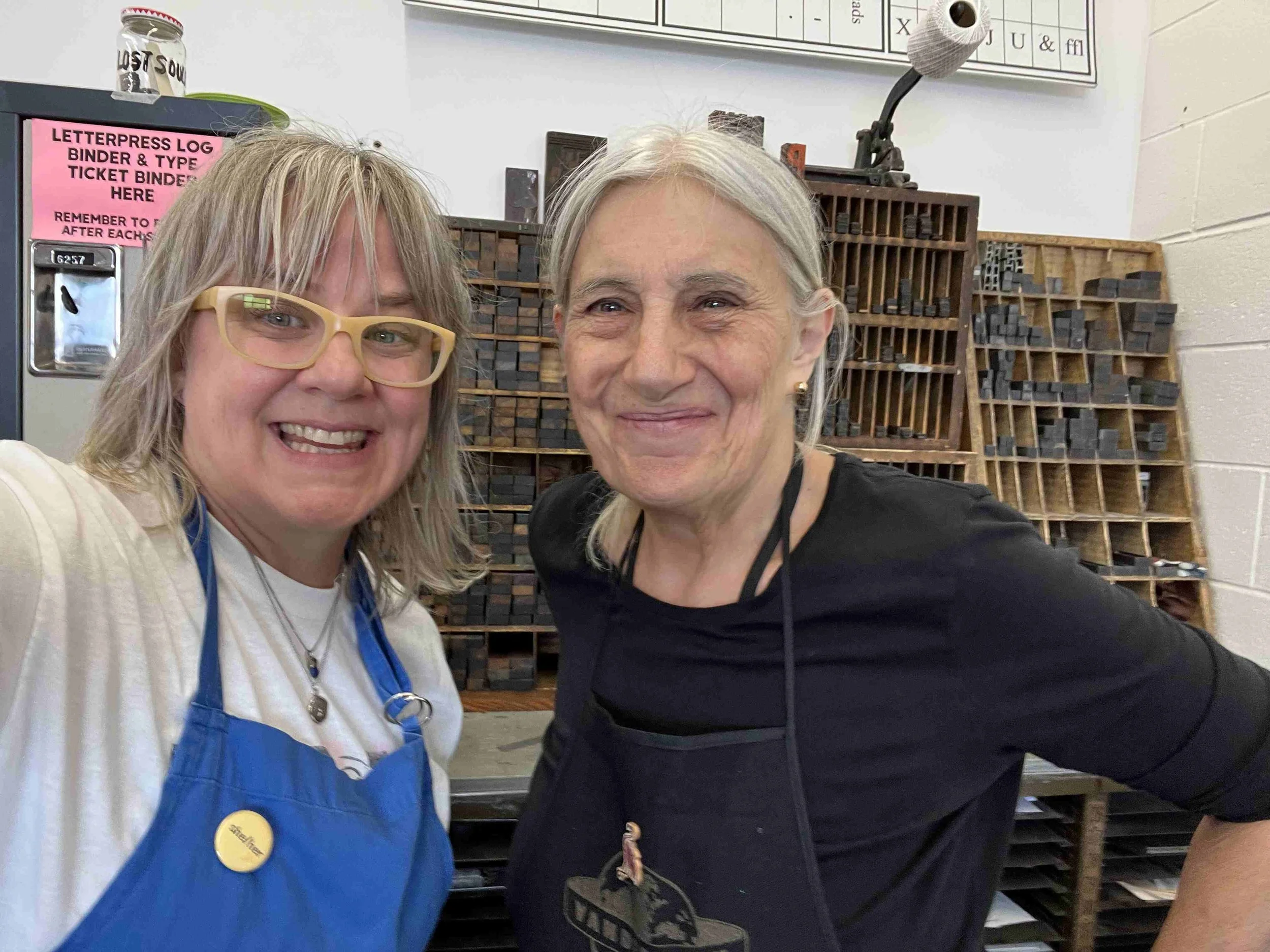 white woman with glasses and shoulder length mousy hair smiles beside white woman with pulled back silver hair in front of letterpress storage shelves