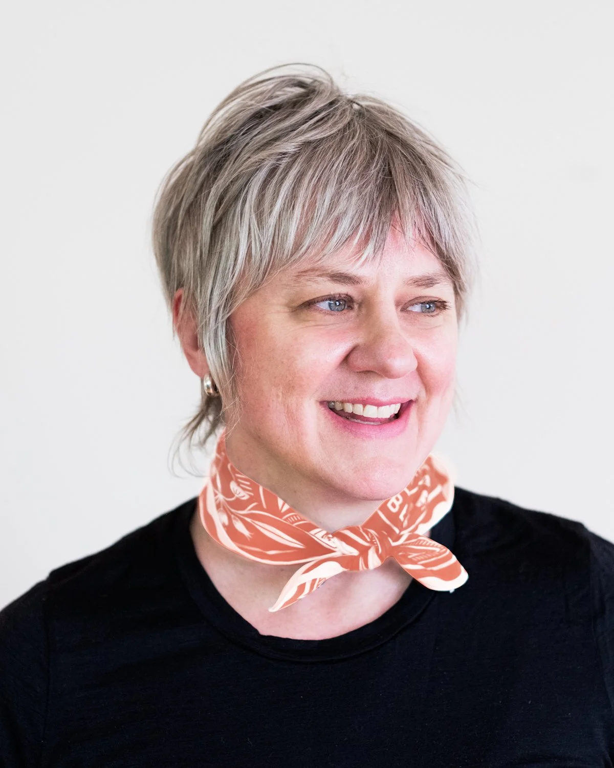 Woman with pale skin and short grey hair wears black shirt and neckerchief in coral and white.