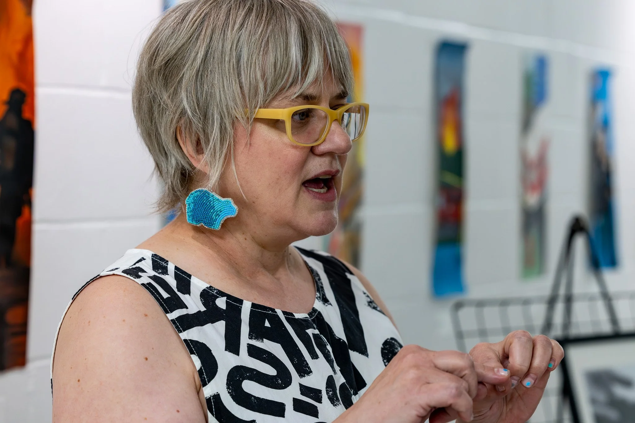 Pale-skinned woman with short, light hair and yellowish glasses wears blue gradient bear-shaped earrings beaded by Wild Mint in Calling Lake and appears to be talking with her mouth and her hands.