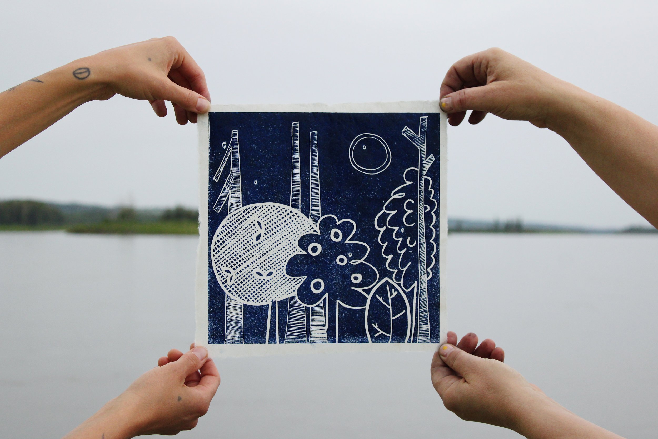 Four light-skinned hands hold paper printed with a sylized treescape design with the Athabasca rivier in the background