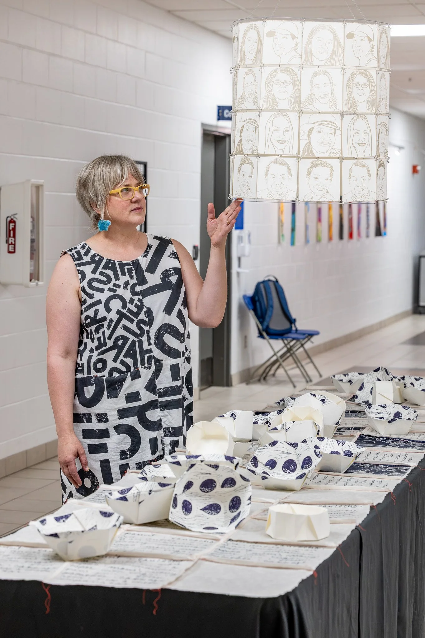 Woman with pale skin, short hair, glasses, and a black and white dress stands beside a table covered in paper cloth and paper folded bowls, with a cylinder above made from paper and printed with the faces of eight people, linked together with wire