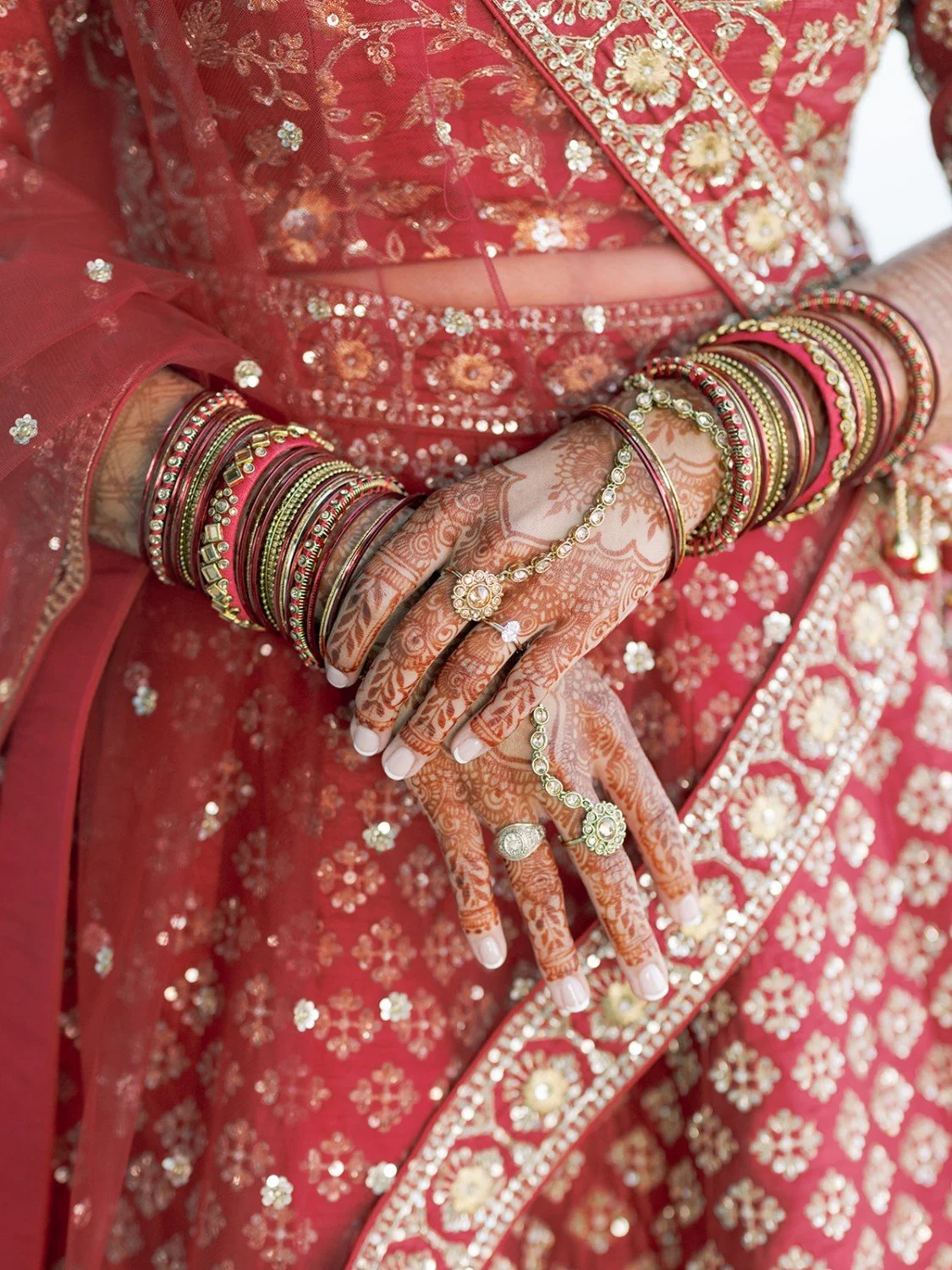 Close-up of a woman's decorated hands with mehndi (henna), adorned with gold and pink bangles, rings, and jewelry, against a richly embroidered red saree with embroidery and sequin details.