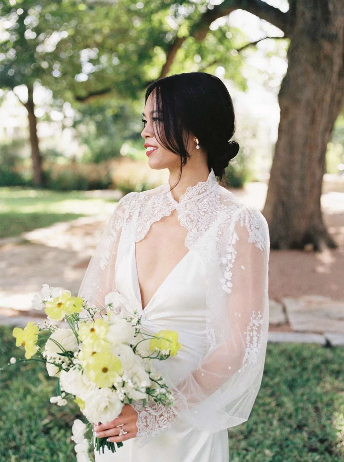 A woman in a white wedding dress holding a bouquet of white and yellow flowers, standing outdoors under a tree with green leaves.