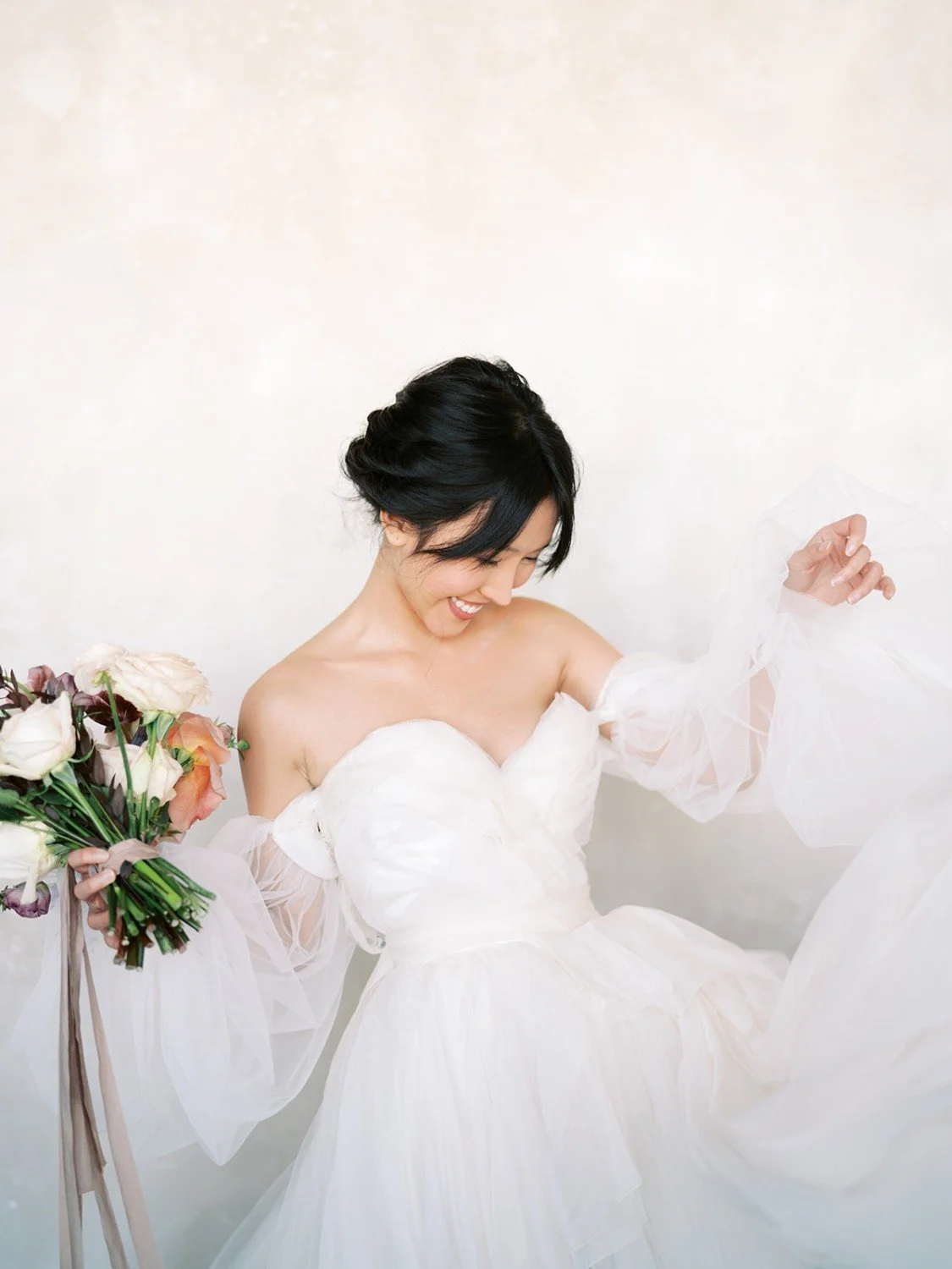 A woman in a white wedding dress holding a bouquet of flowers, smiling and looking down.