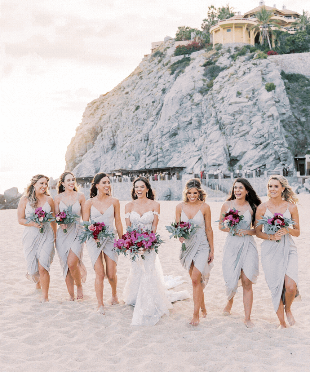 A bride in a white wedding dress walking on a beach with seven bridesmaids in light gray dresses, all holding bouquets of pink and purple flowers, with large rocks and houses on a cliff in the background.