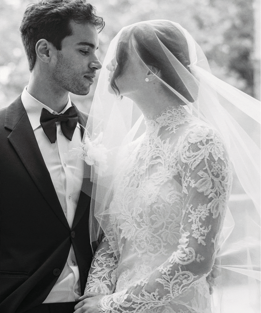 A black-and-white photo of a bride and groom standing close with foreheads touching, in wedding attire, outdoors.