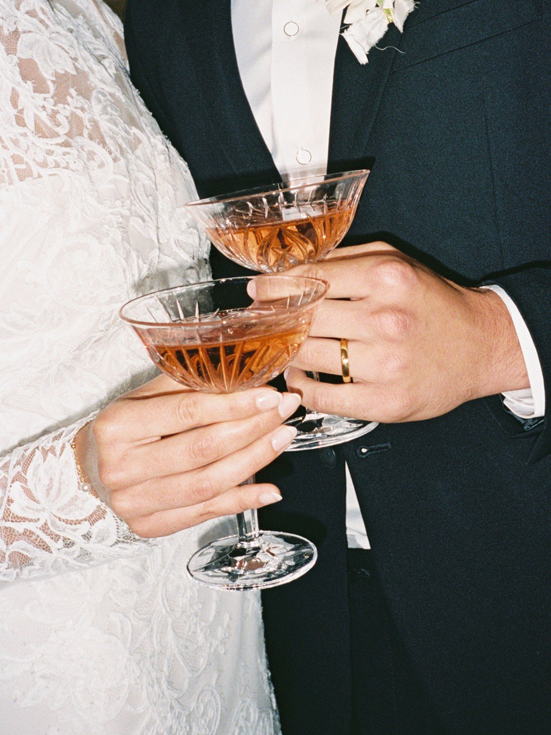 A couple dressed in wedding attire holding crystal cocktail glasses with pinkish-orange drinks, celebrating at their wedding.