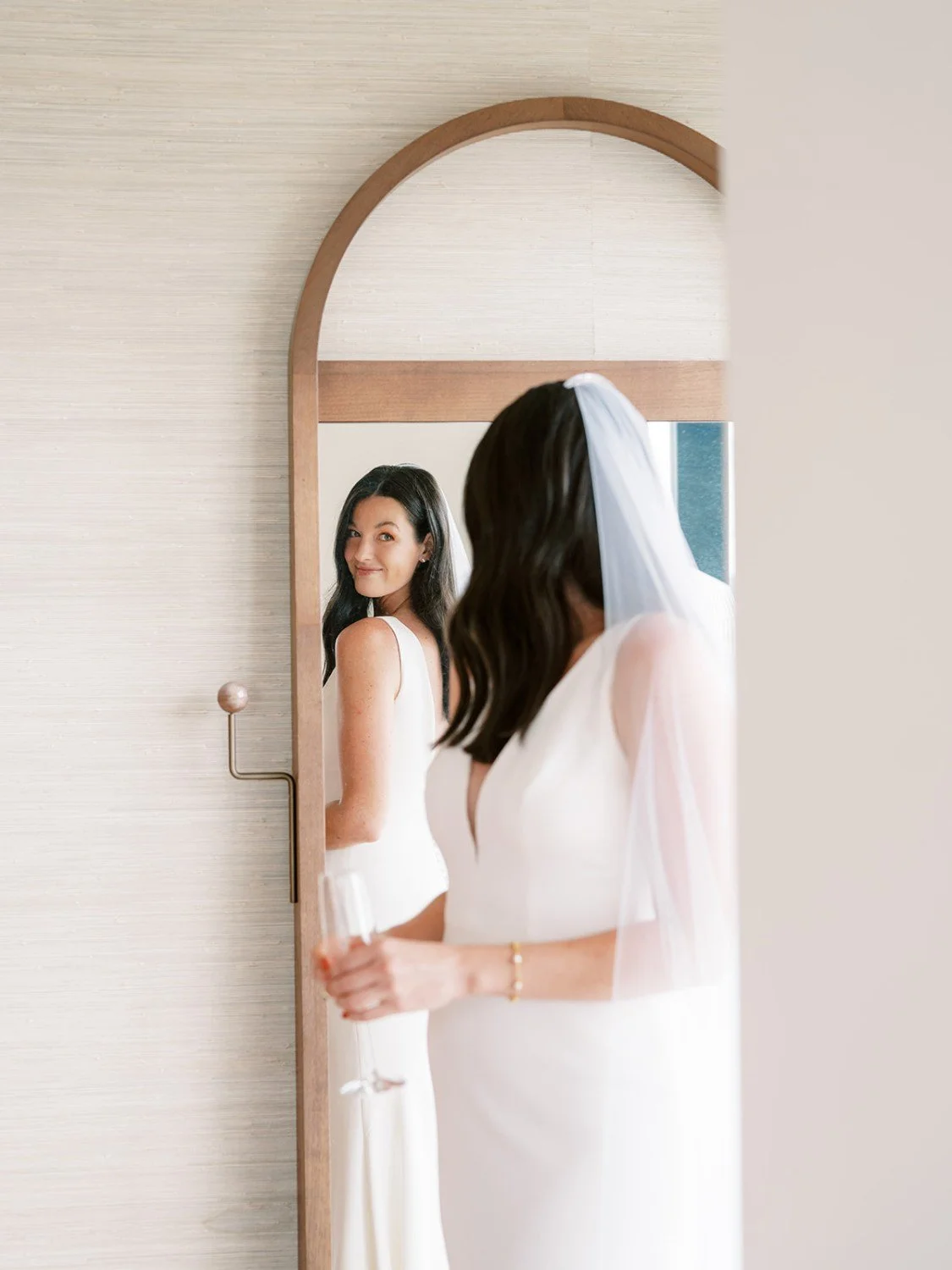 Bride with dark wavy hair and white wedding dress holding a glass, looking at herself in a mirror with another woman reflected.