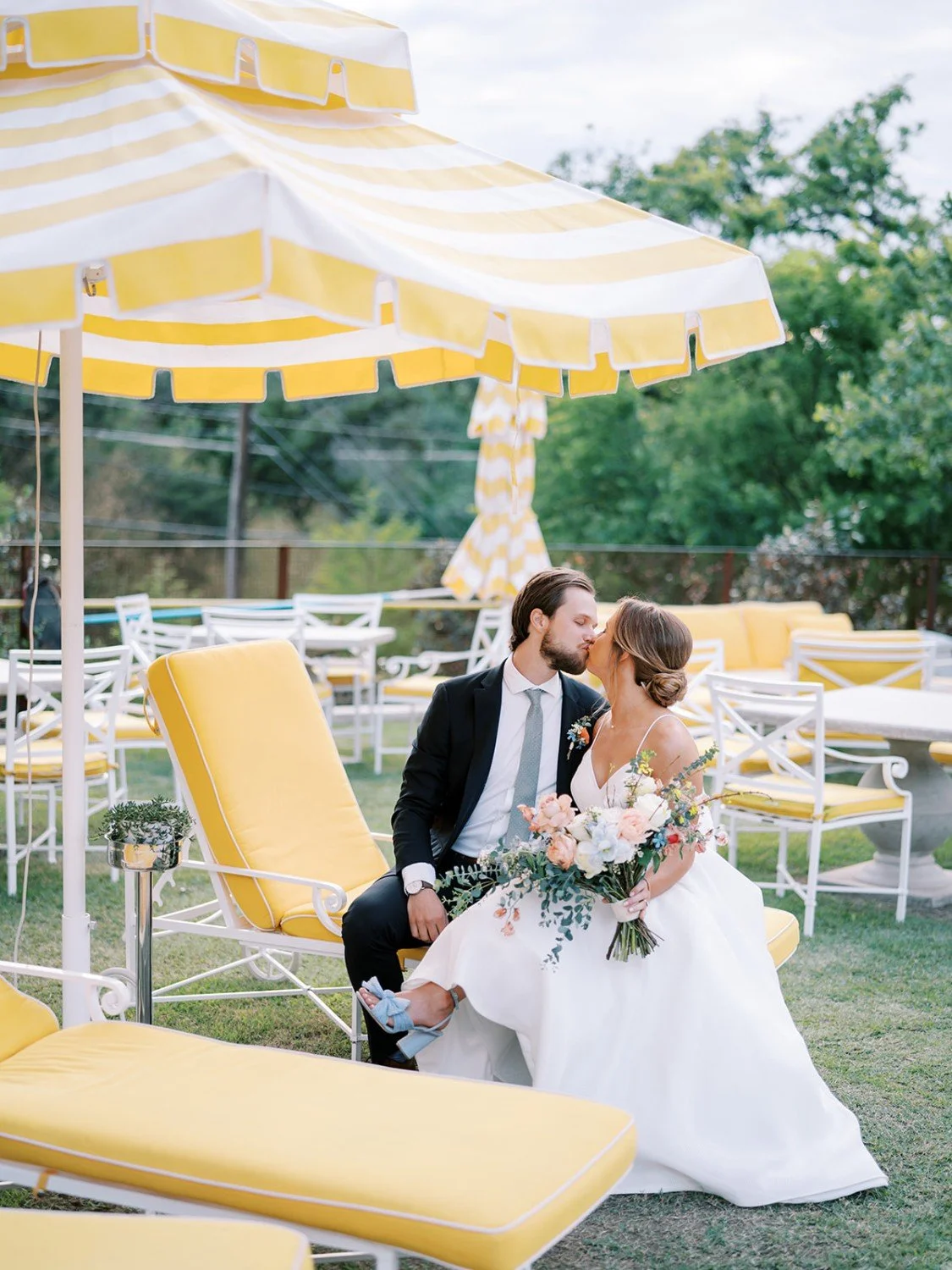 A bride and groom sharing a kiss outdoors on a sunny day, surrounded by yellow umbrellas and patio furniture, with the bride holding a bouquet of flowers.