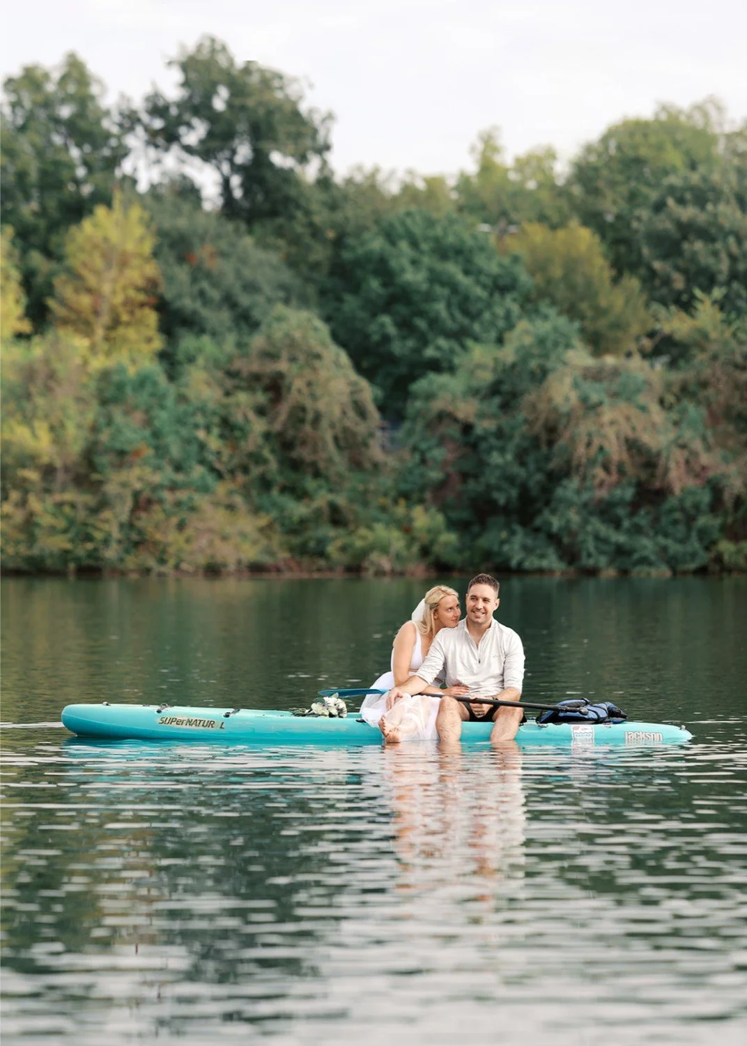 A couple sitting on a paddleboard in a lake, surrounded by green trees.