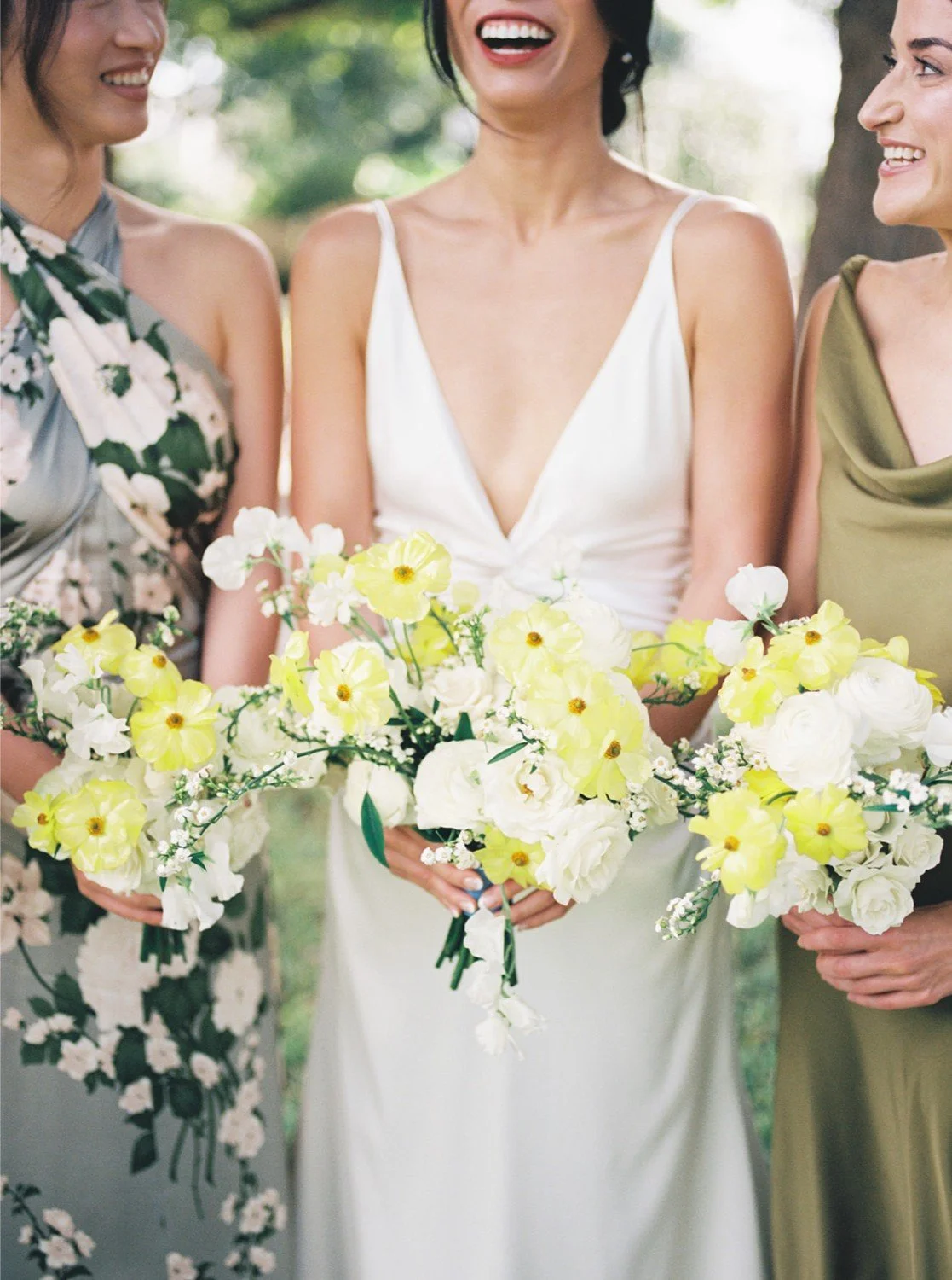 Three women dressed in floral, white, and olive green dresses, holding bouquets of white and yellow flowers, standing outdoors.
