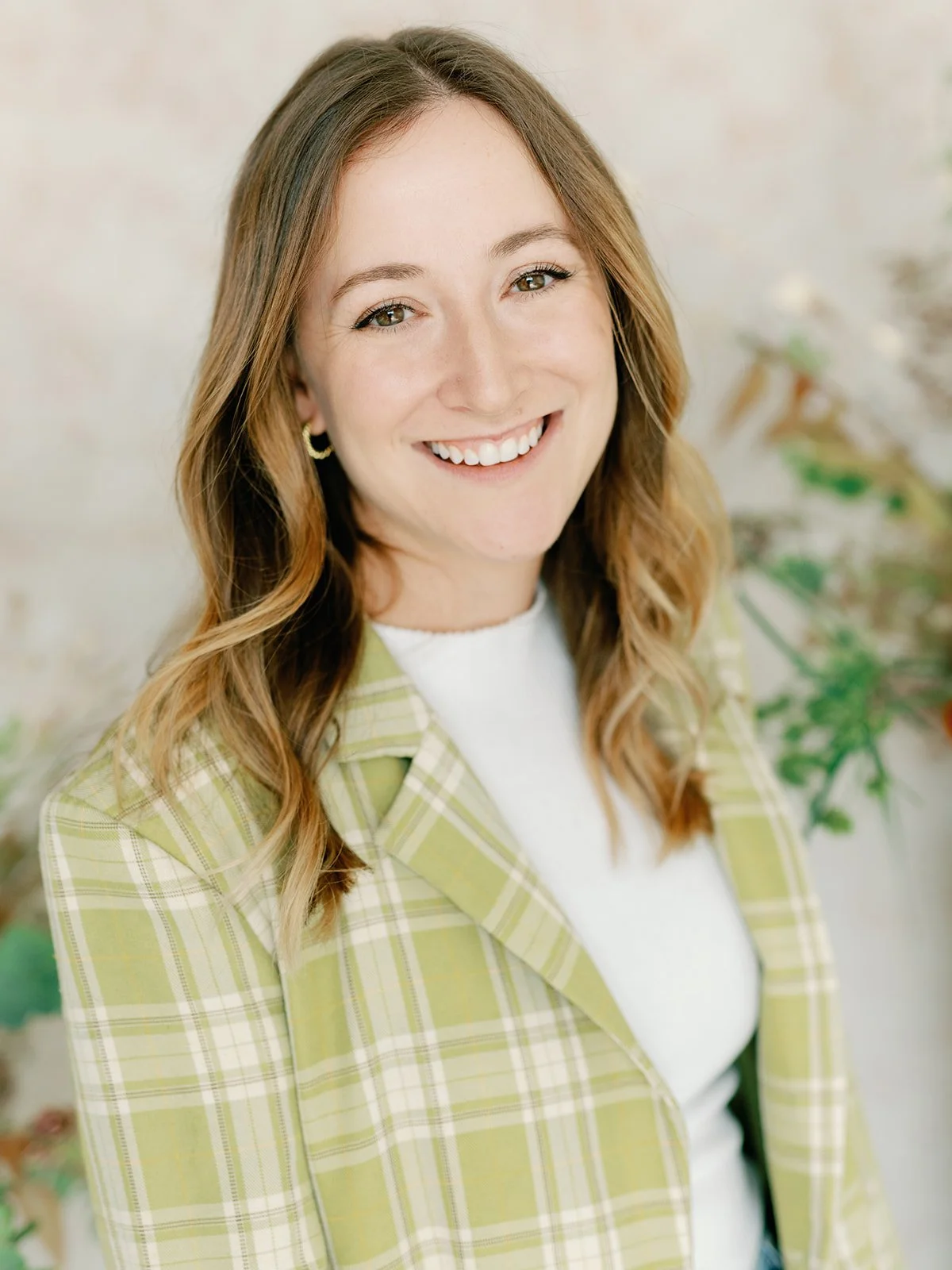 A smiling woman with long, wavy brown hair, wearing a light green plaid blazer and a white top, standing in front of a blurred background with plants.