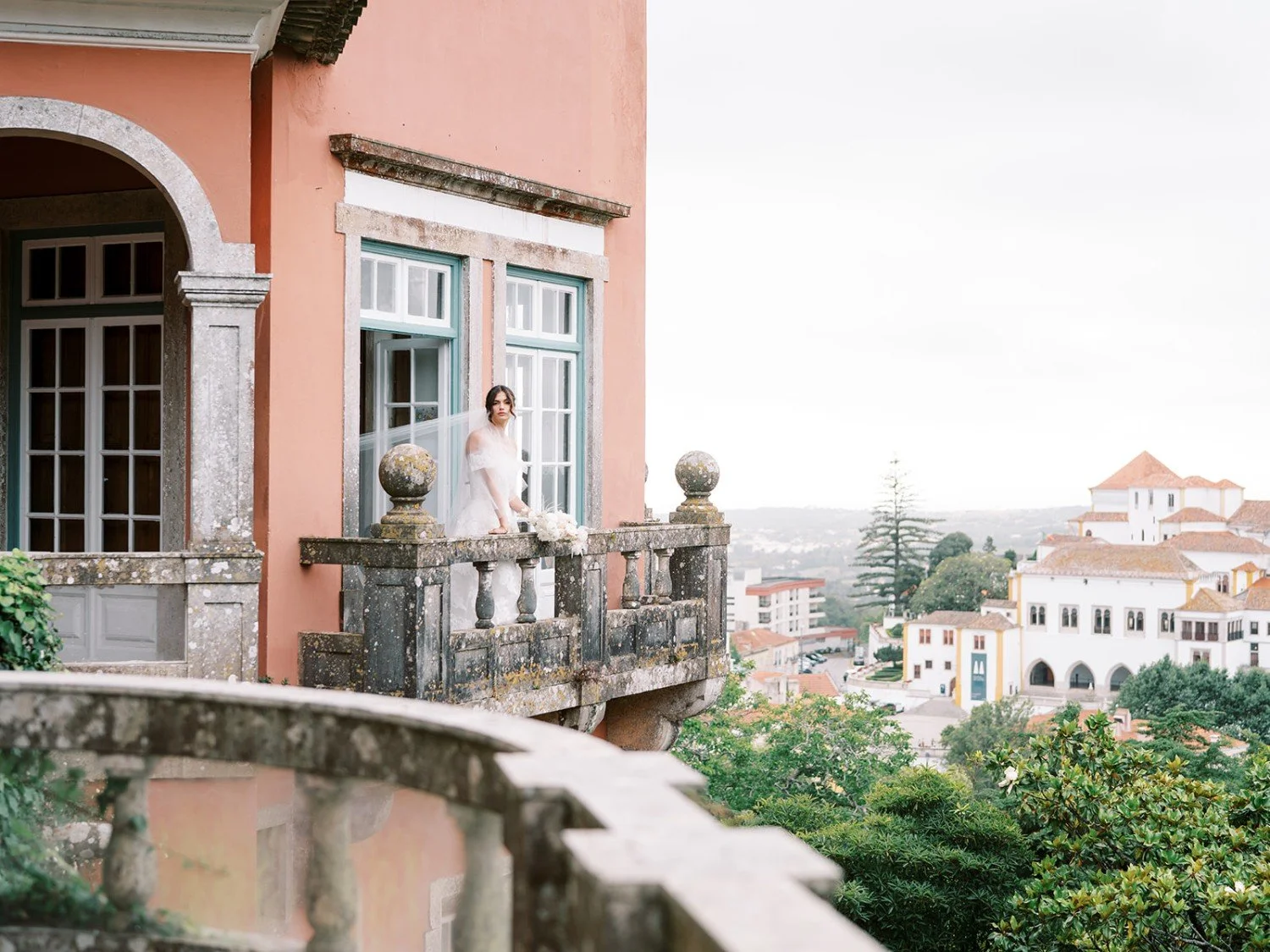 A woman in a white dress standing on a balcony of a pink building with a view of a town with red-tiled roofs and white buildings in the distance.