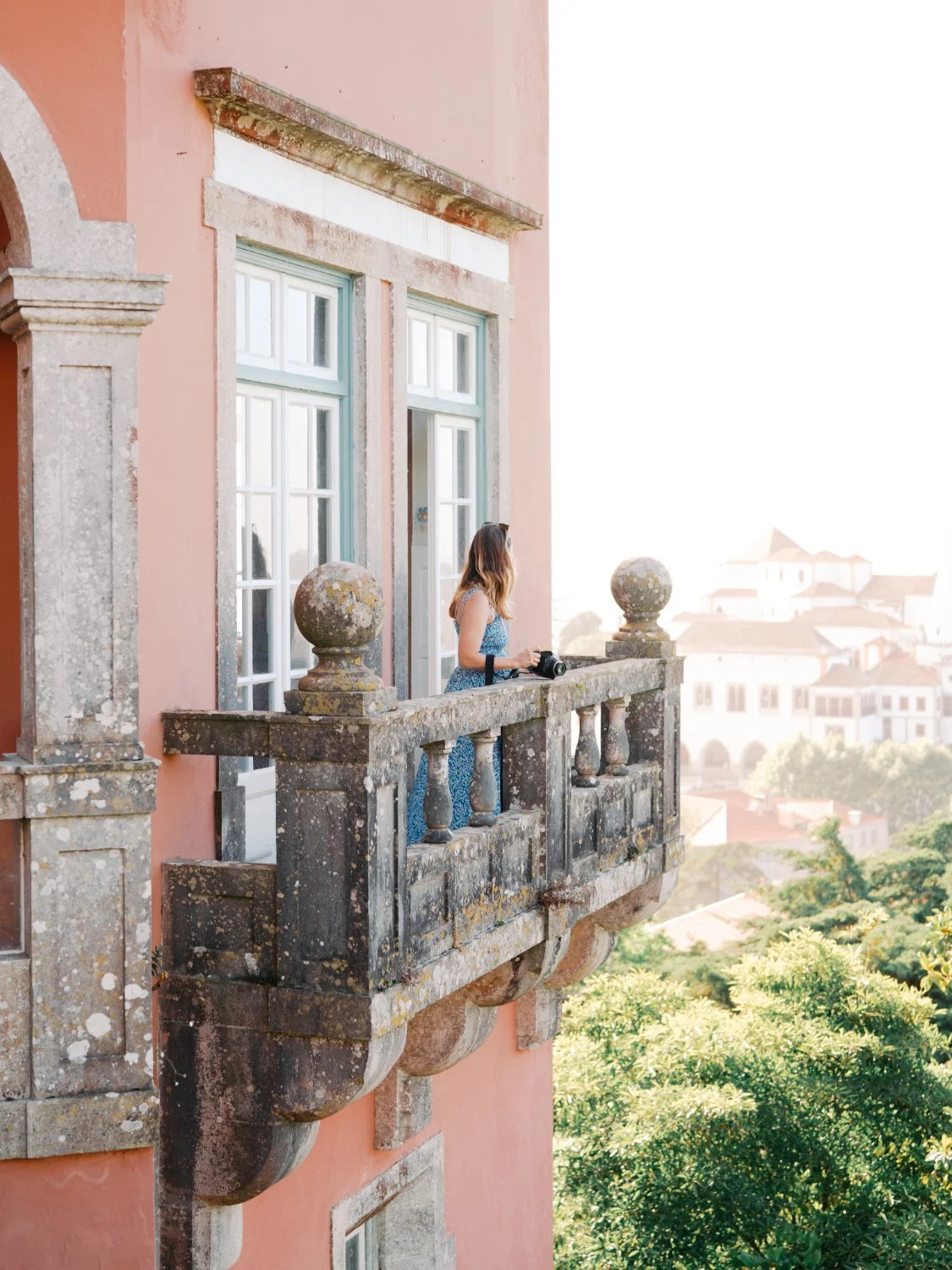 Woman standing on a stone balcony of a historic building with pink walls, holding a camera, overlooking a cityscape with lush green trees and old buildings in the background.