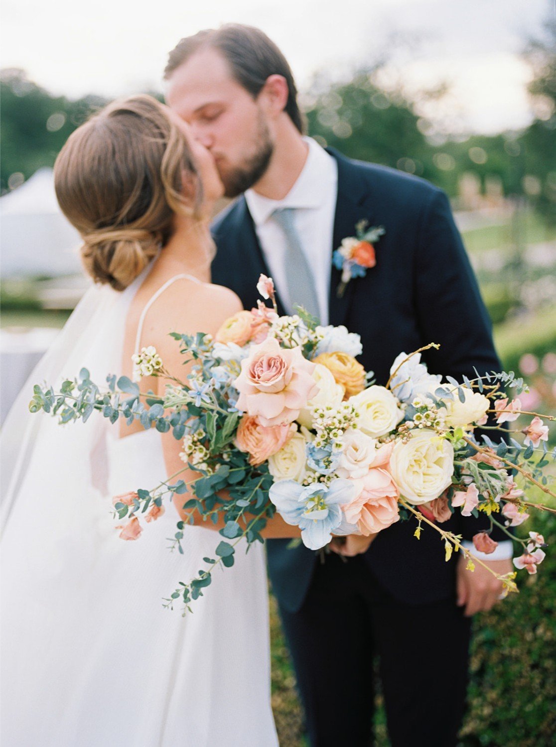 A bride and groom sharing a kiss outdoors during their wedding, with the bride holding a bouquet of pastel-colored flowers, including roses and greenery.