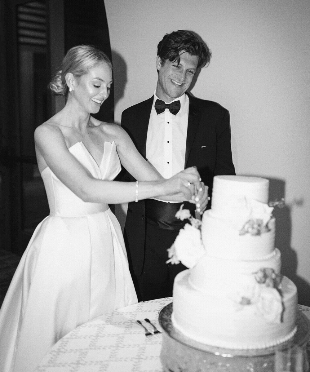A bride and groom in wedding attire cutting a wedding cake together, both smiling, in black and white.