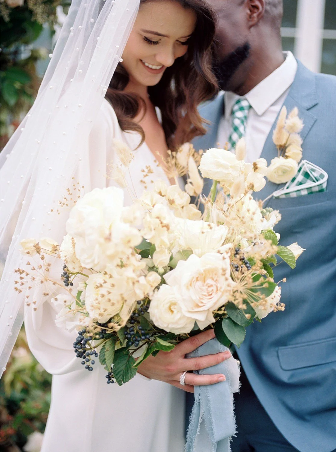 A wedding couple close together, smiling, with the bride holding a bouquet of white and cream-colored flowers, and the groom in a light blue suit with a patterned green tie and pocket square.