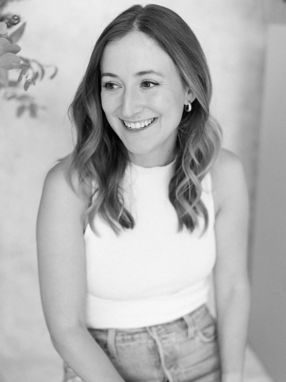 A woman with wavy hair smiling while looking off to the side, wearing a sleeveless top and jeans, with a blurred background and some leaves on the left side.