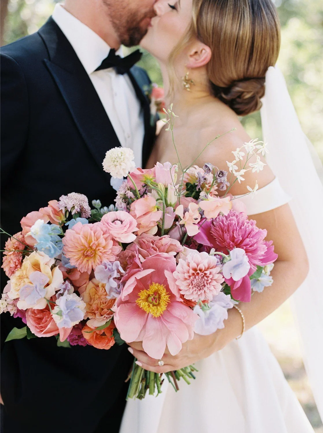 A bride and groom sharing a kiss on their wedding day, with the bride holding a large, colorful bouquet of pink, peach, purple, and blue flowers.