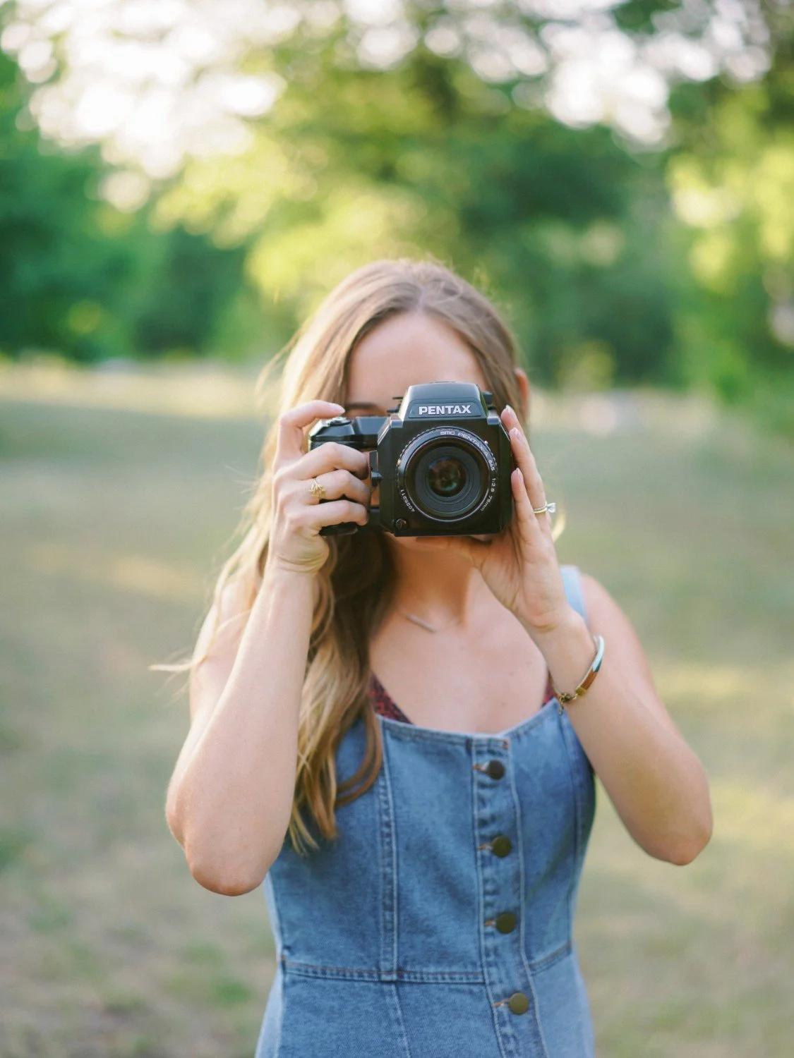 A woman holding a Pentax camera, taking a photo in an outdoor park with green trees in the background.