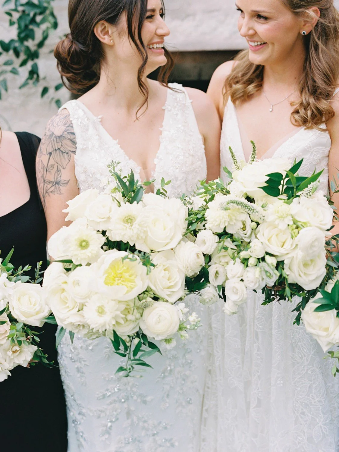Two women in wedding dresses smiling and holding bouquets of white flowers with green leaves.