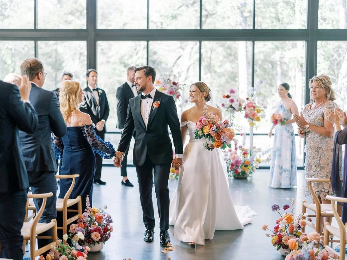A bride and groom walking hand in hand down the aisle during their wedding ceremony, smiling and surrounded by guests clapping in a brightly lit, flower-filled venue with large windows.
