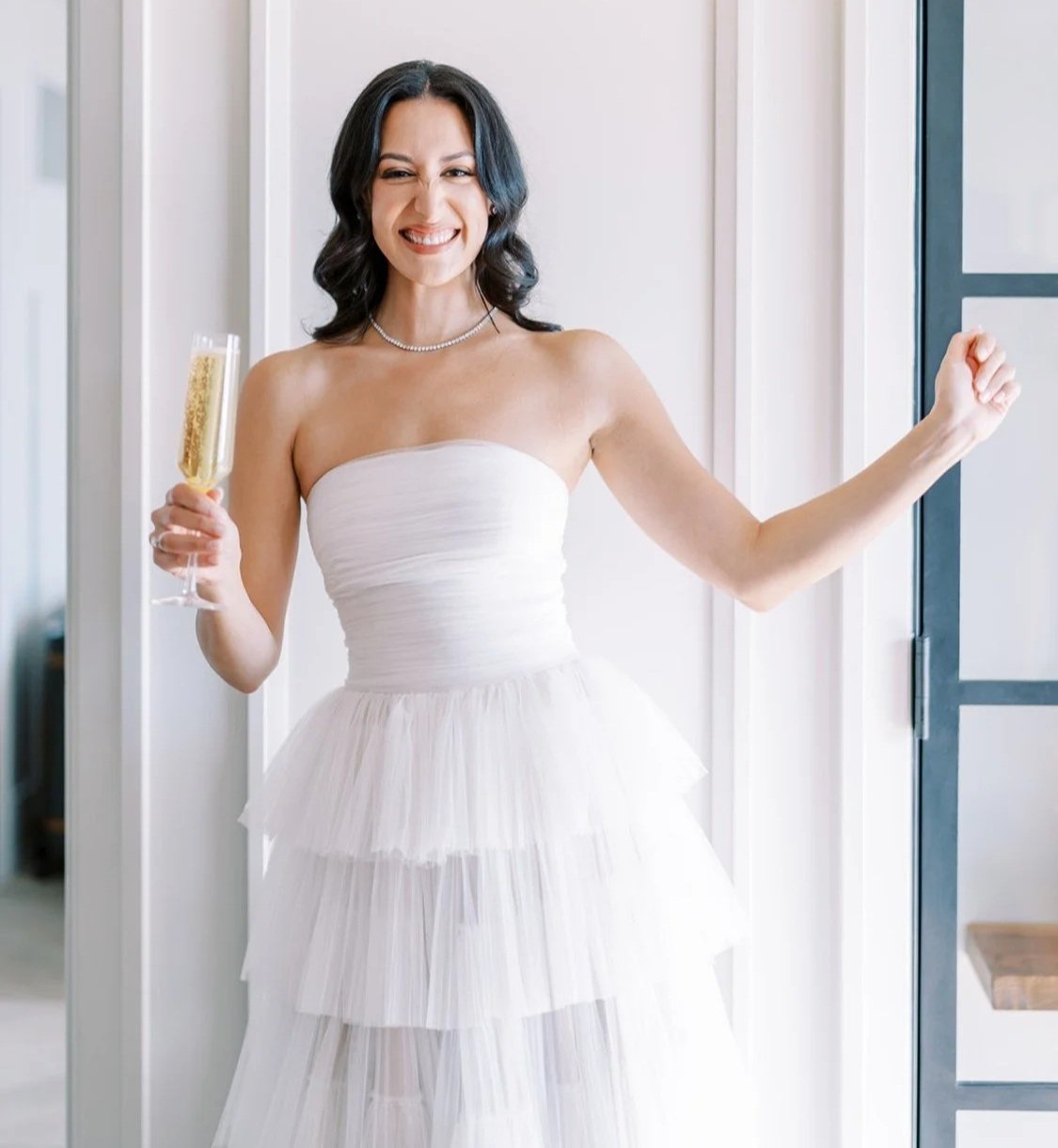 A woman in a strapless white dress holding a glass of champagne, smiling and making a confident gesture.