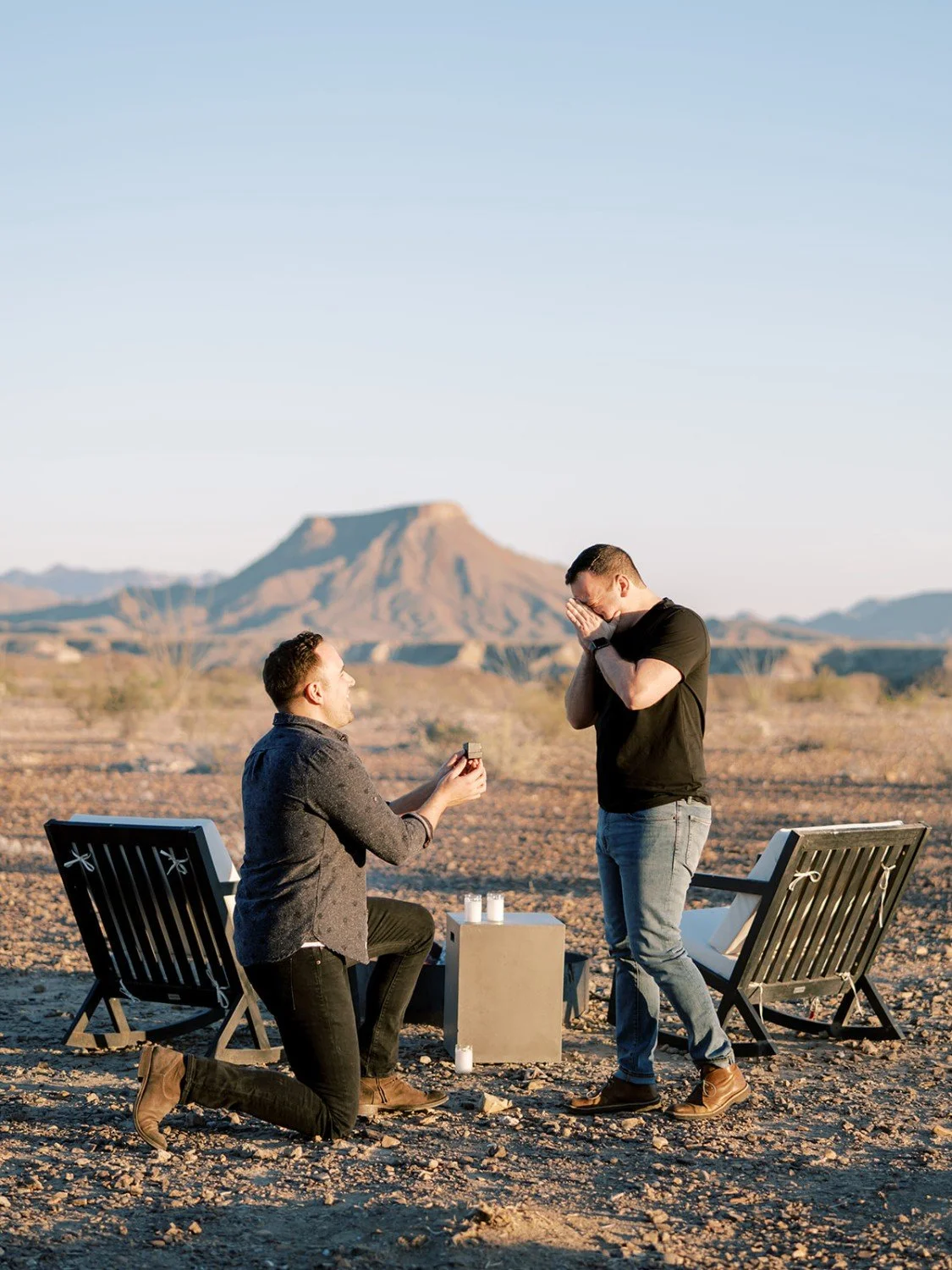A man on one knee holding a ring, proposing to another man in a desert landscape with mountains in the background, during sunset.