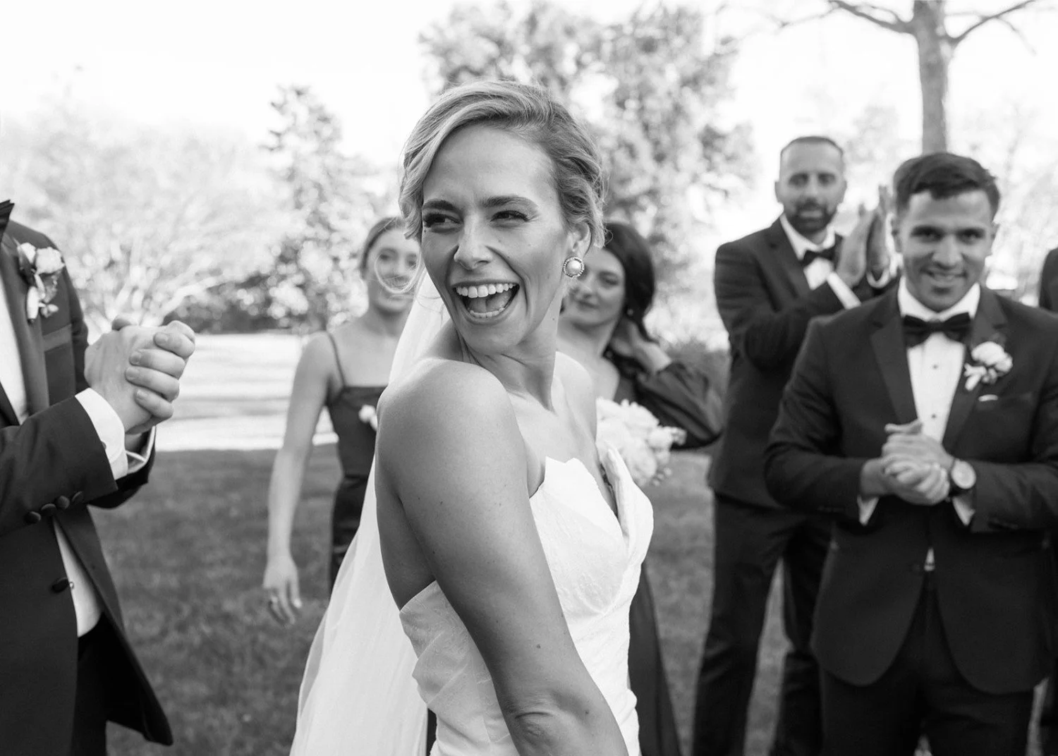 Happy bride in wedding dress laughing and smiling during outdoor wedding celebration with bridal party in the background.
