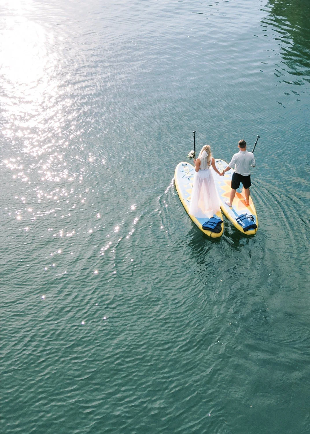A couple in wedding attire paddleboarding on the water, holding hands.