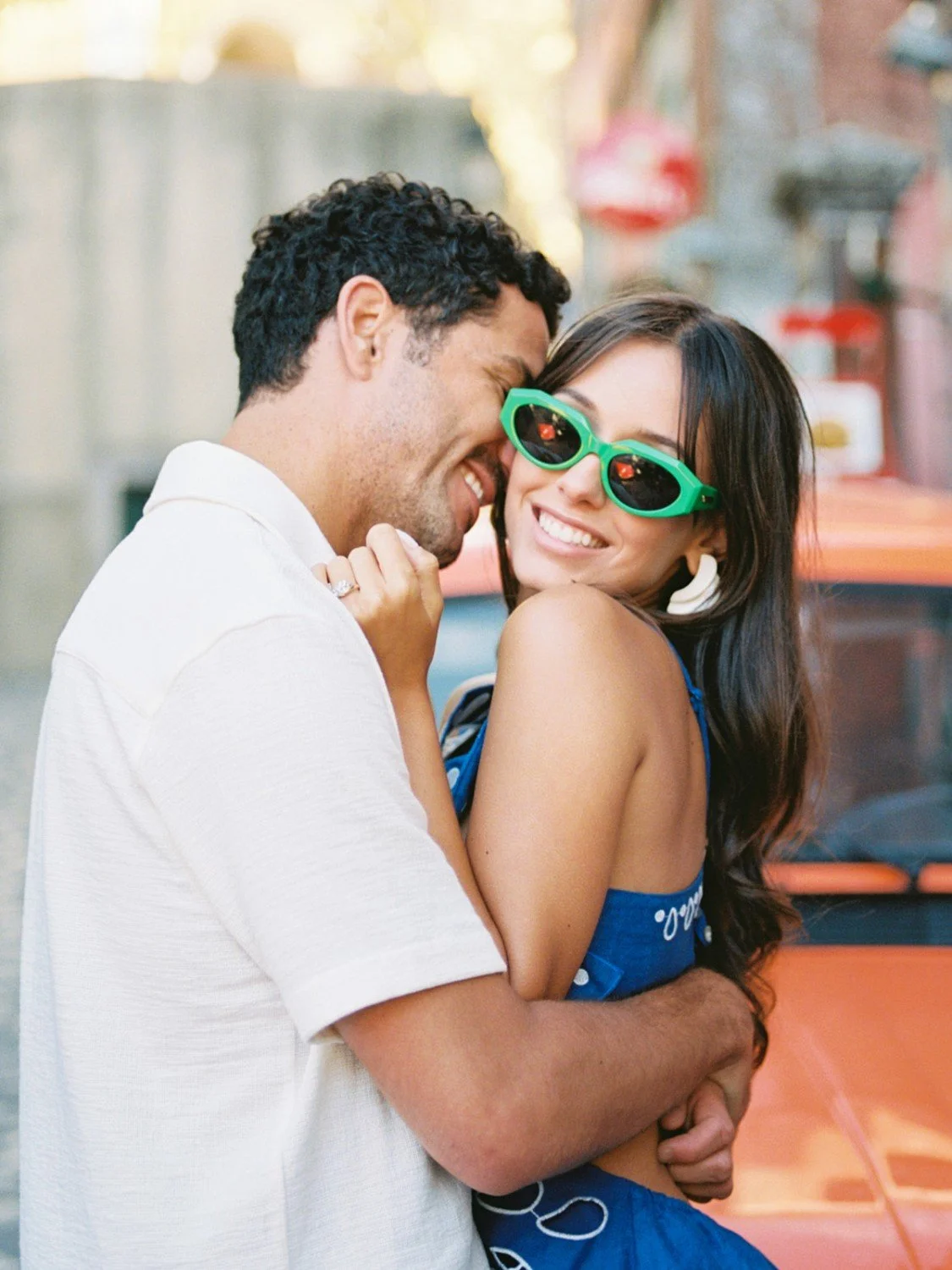 A happy couple embracing outdoors, smiling, with the woman wearing green sunglasses and a blue top, and a vintage car in the background.