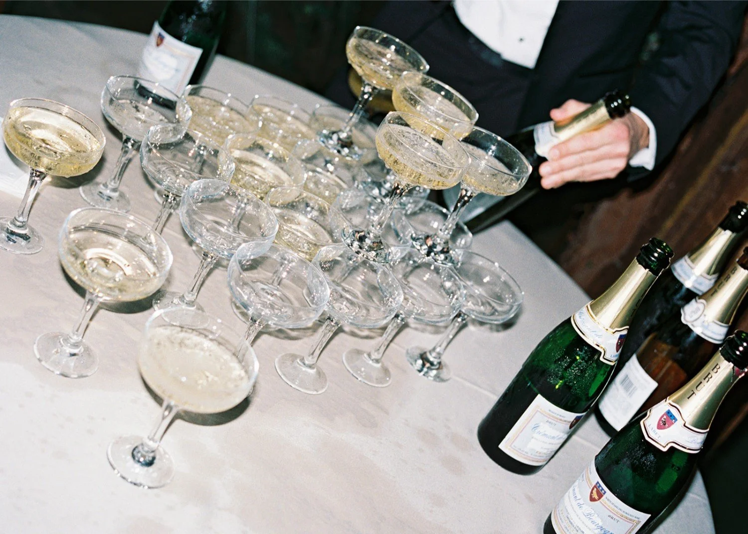 A table with several champagne glasses filled with sparkling wine and bottles of champagne, with a person in a dark suit pouring or handling the bottles.