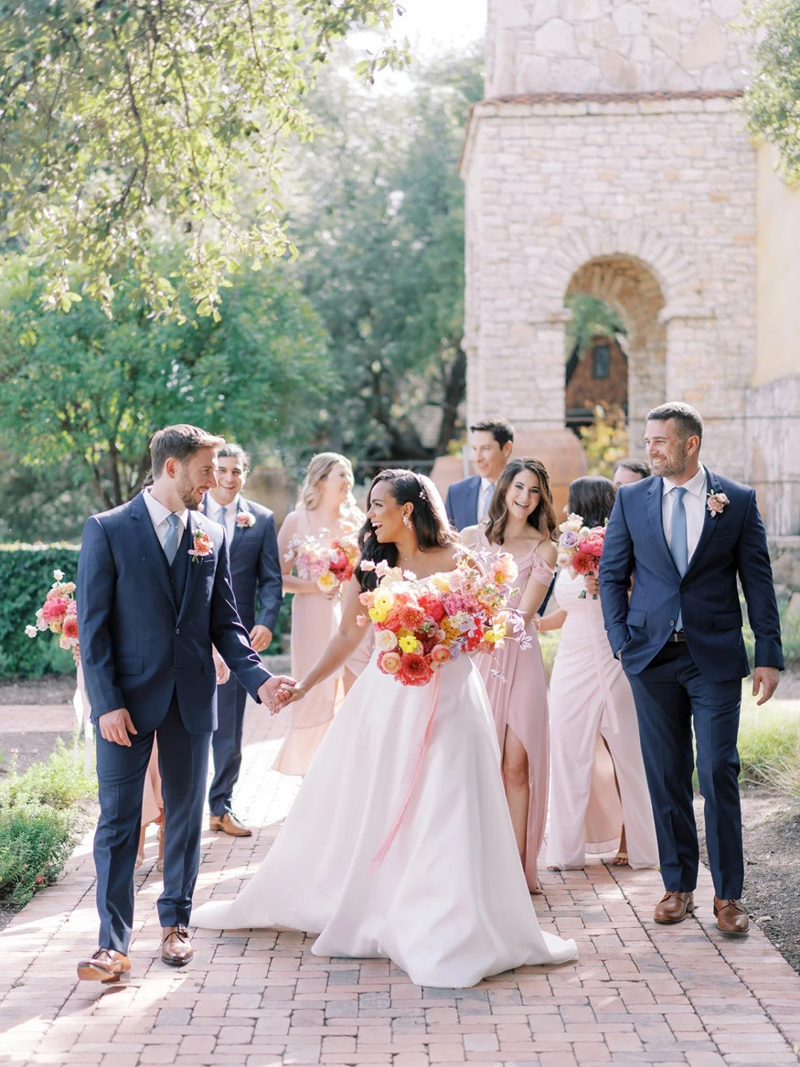 A group of people dressed in wedding attire walking outdoors on a brick path, carrying bouquets of colorful flowers and smiling.