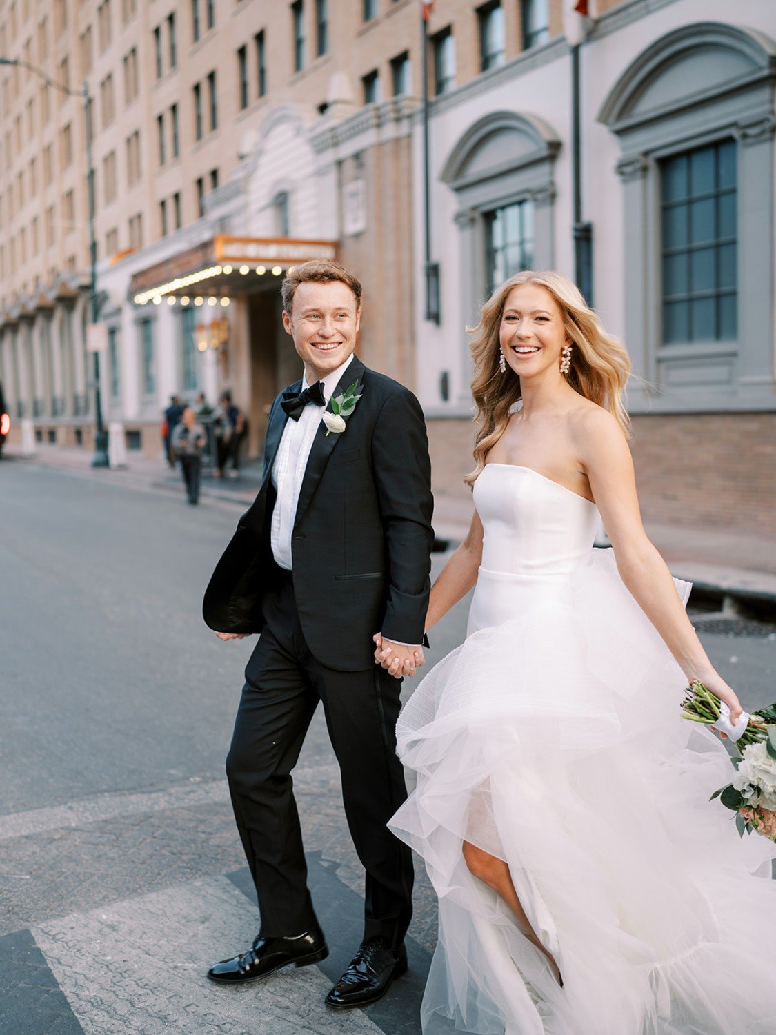 A newlywed couple, the groom in a black tuxedo and the bride in a white wedding gown, holding hands and walking on a city street. The bride is holding a bouquet of flowers, and both are smiling.