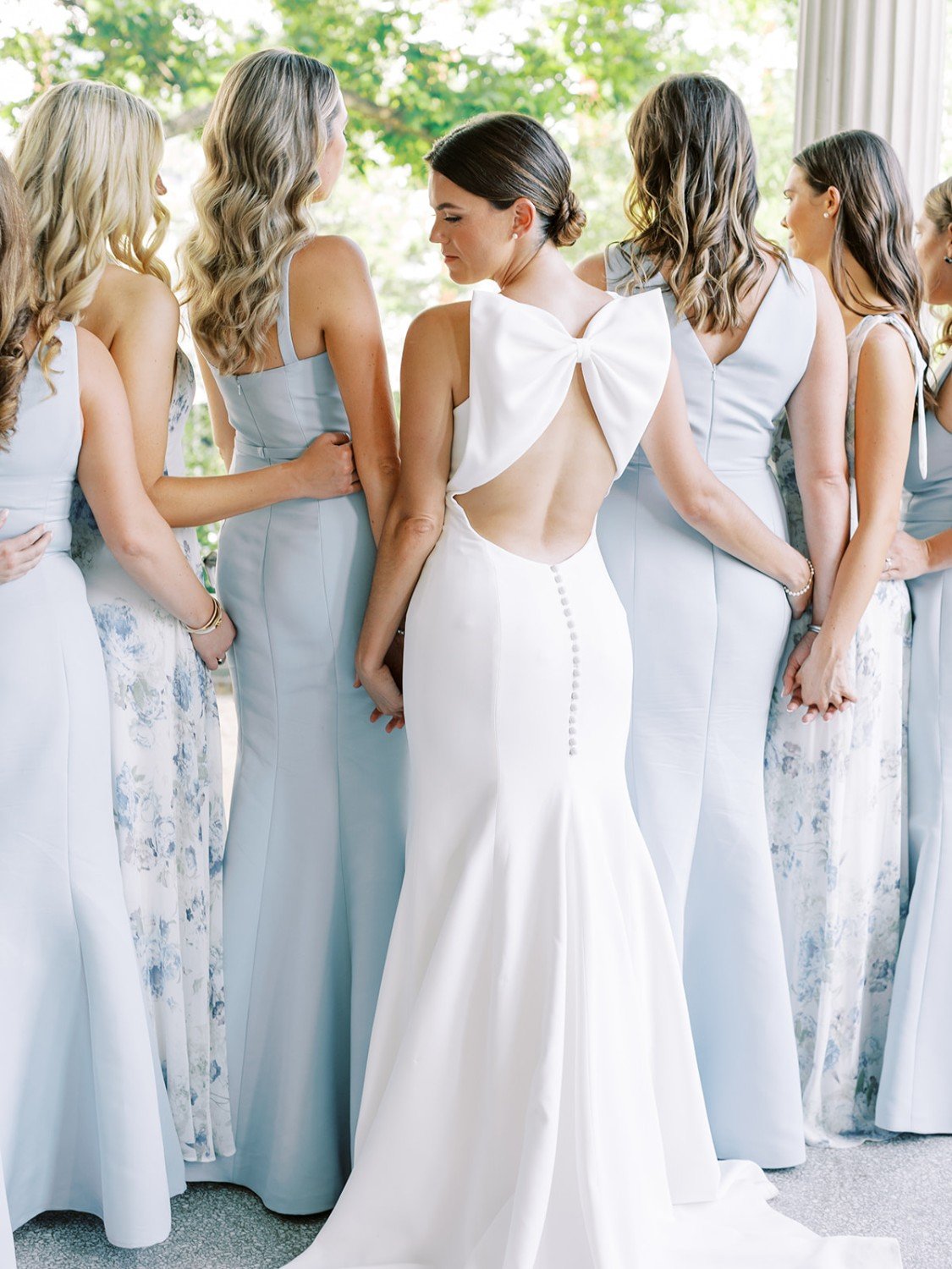 Bride in a white gown with an open back and large bow detail, surrounded by bridesmaids in light blue dresses, during a wedding preparation.