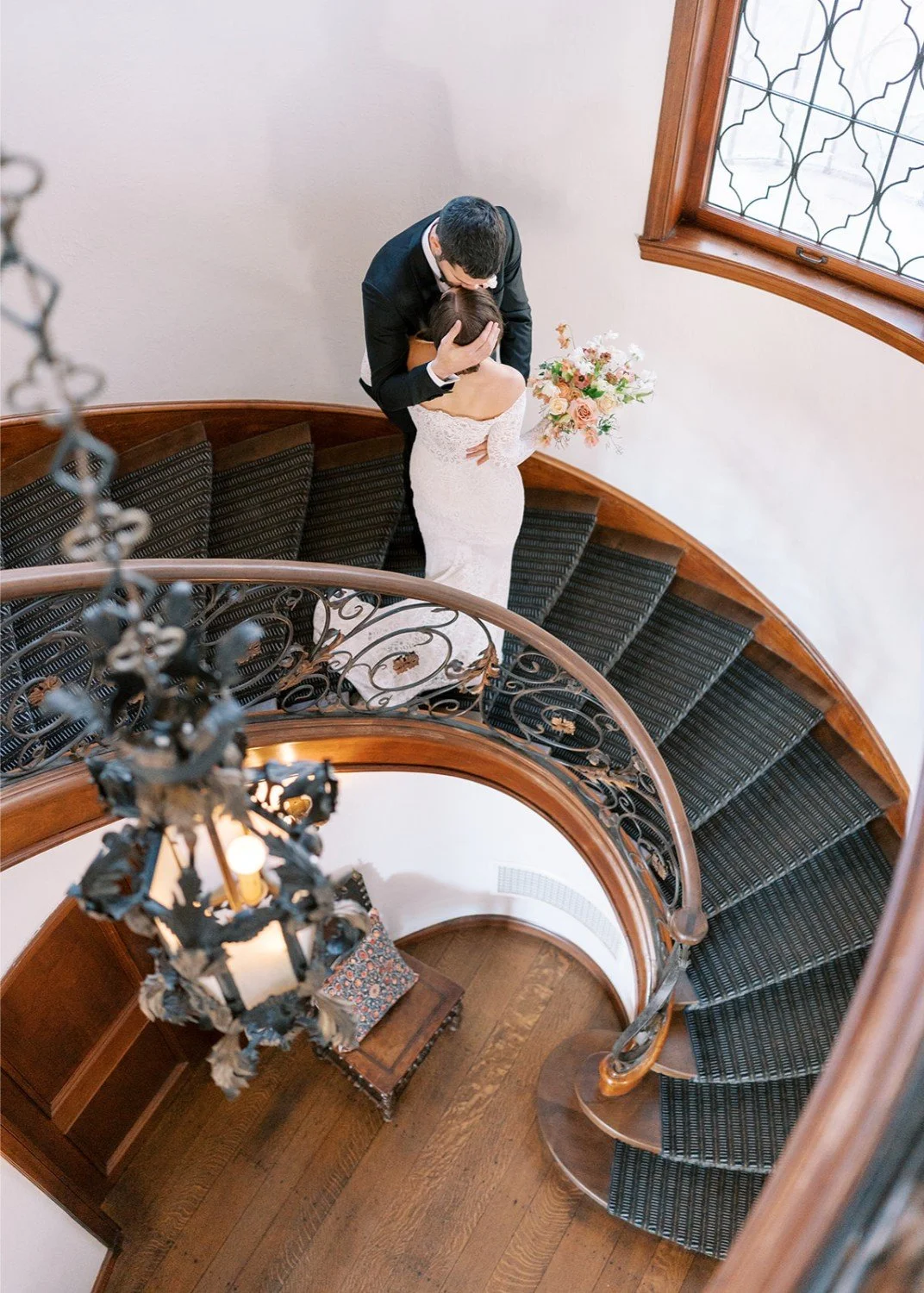 A couple standing on a curved staircase, with the man kissing the woman's forehead. The woman is in a white wedding dress holding a bouquet, and the man is in a black suit. The photo is taken from above, showing a chandelier hanging from the ceiling and a window on the wall.