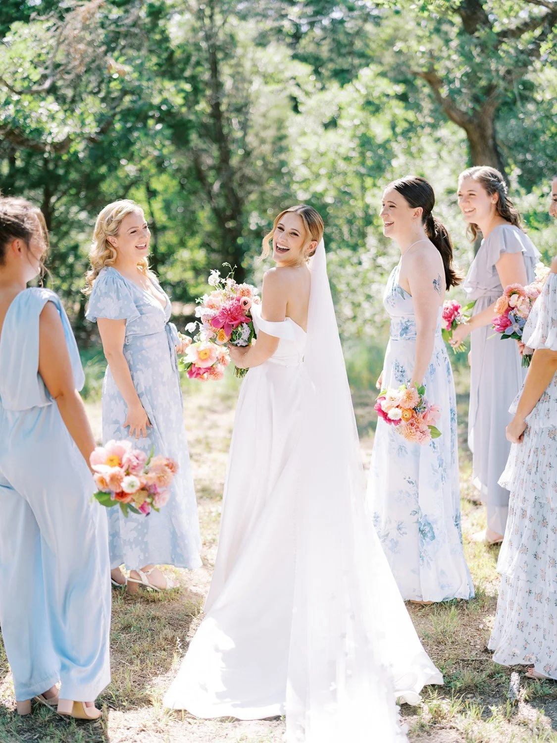 A bride in a white wedding dress holding a bouquet of pink and white flowers stands outdoors in a sunlit forested area, surrounded by six women in pastel-colored dresses holding similar bouquets, all smiling and looking at each other.