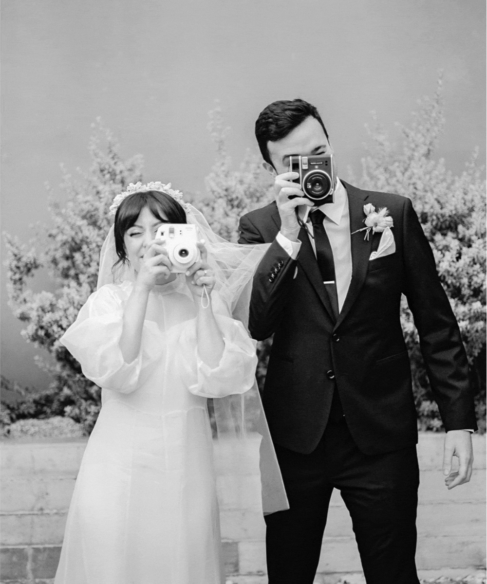 A bride and groom taking a photo with vintage cameras on their wedding day, standing in front of a floral backdrop.