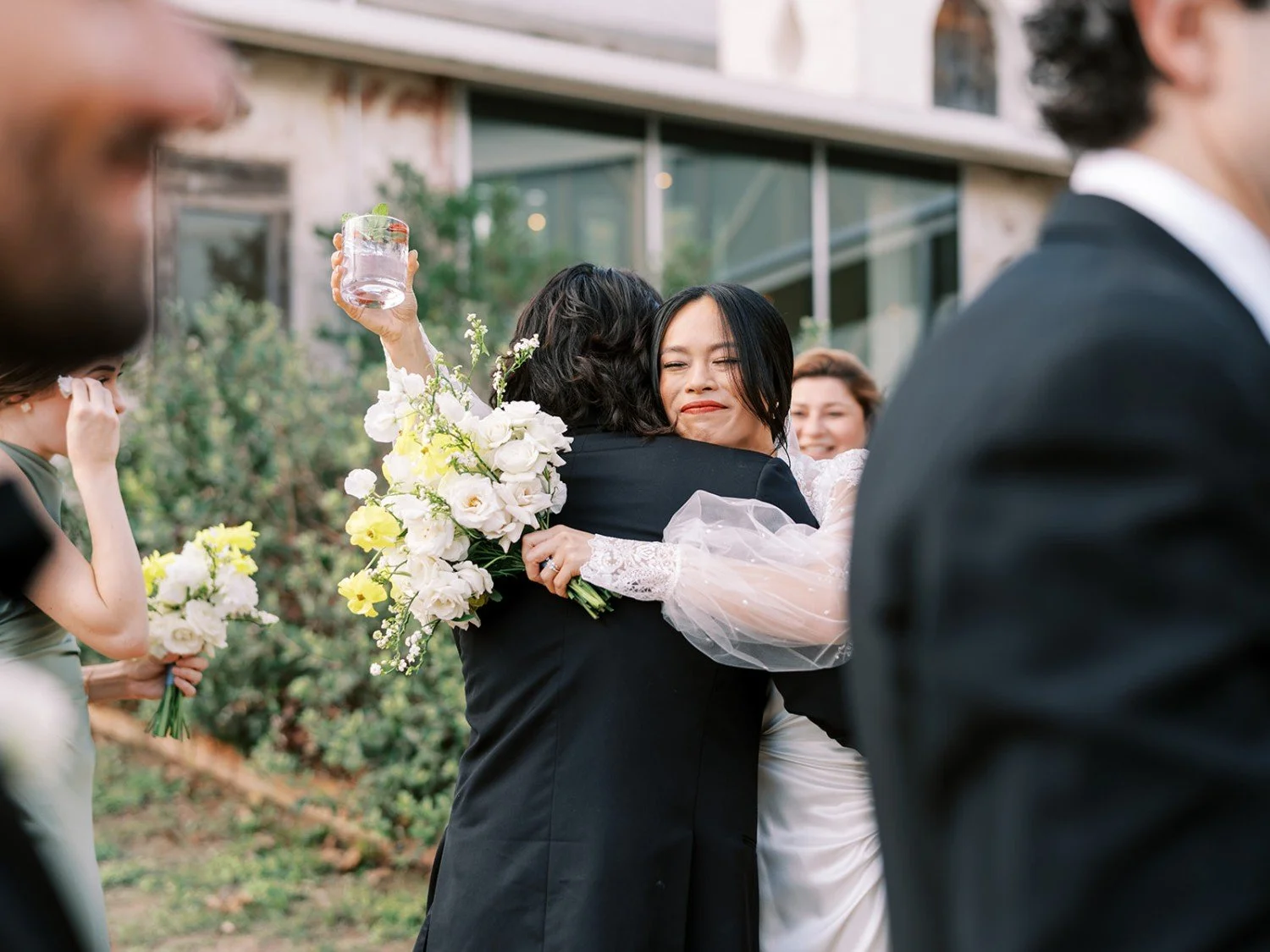 Two women hugging at a wedding, one with a bouquet of white flowers and a big smile, the other holding a glass of drink, surrounded by guests.