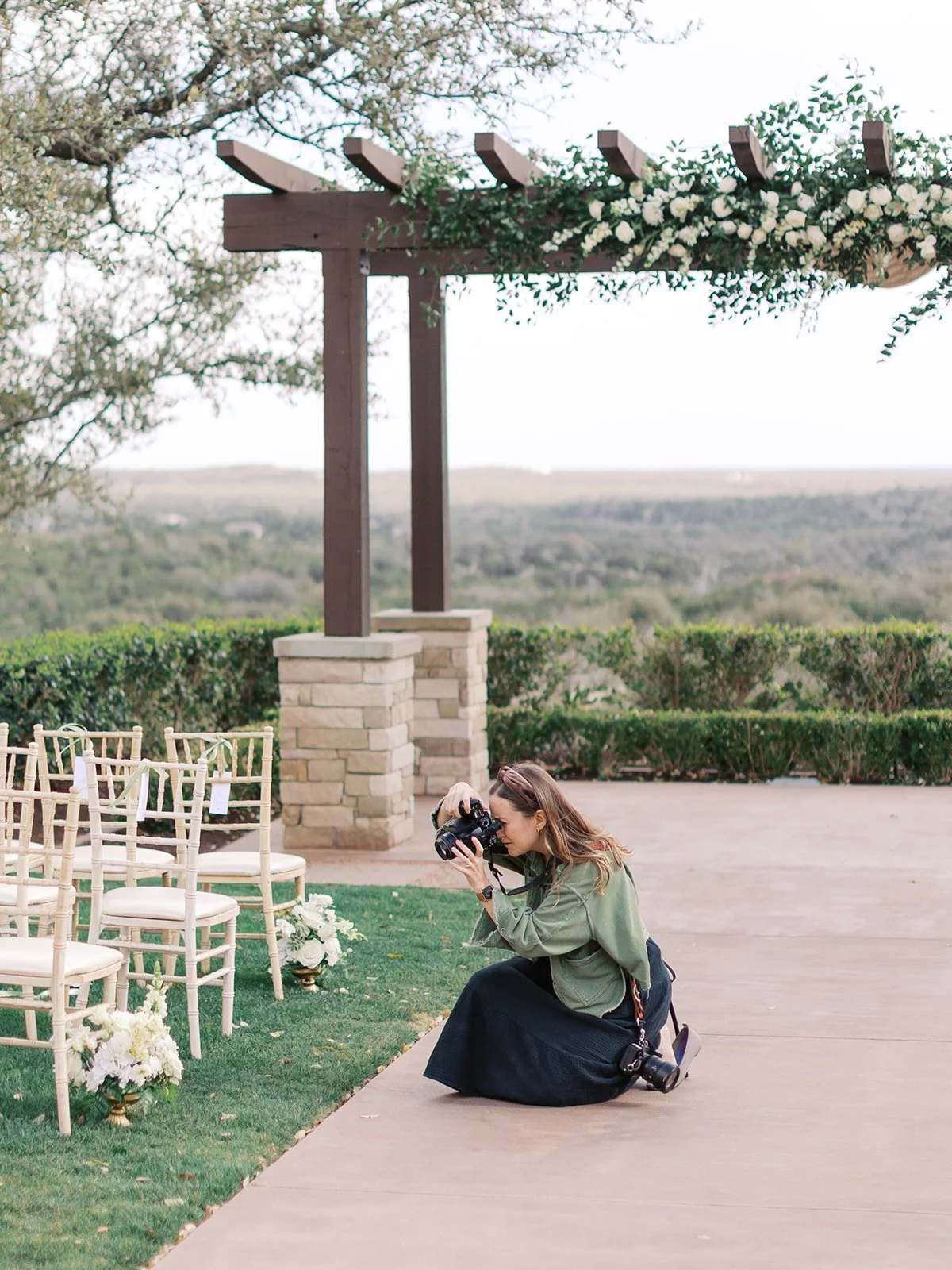 A woman kneeling on the ground taking photos with a camera at an outdoor event setup, with chairs and floral arrangements visible, under a wooden pergola decorated with flowers.