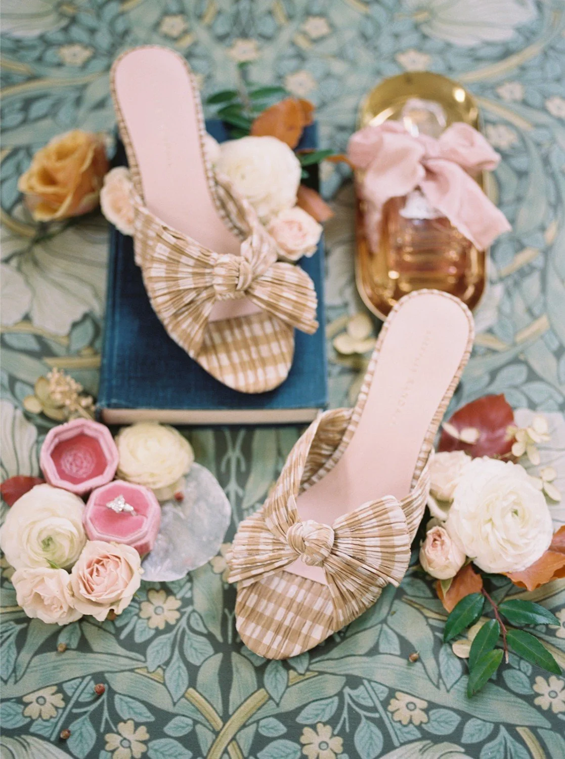 A pair of beige high-heeled shoes with pleated bows on the toes, surrounded by flowers, perfume bottles, and jewelry on a patterned tablecloth.