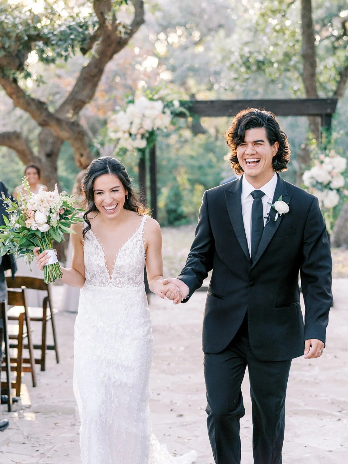 A newlywed couple walking joyfully outdoors, holding hands, with the bride carrying a bouquet of flowers, surrounded by wedding decorations and guests, in a wooded setting.