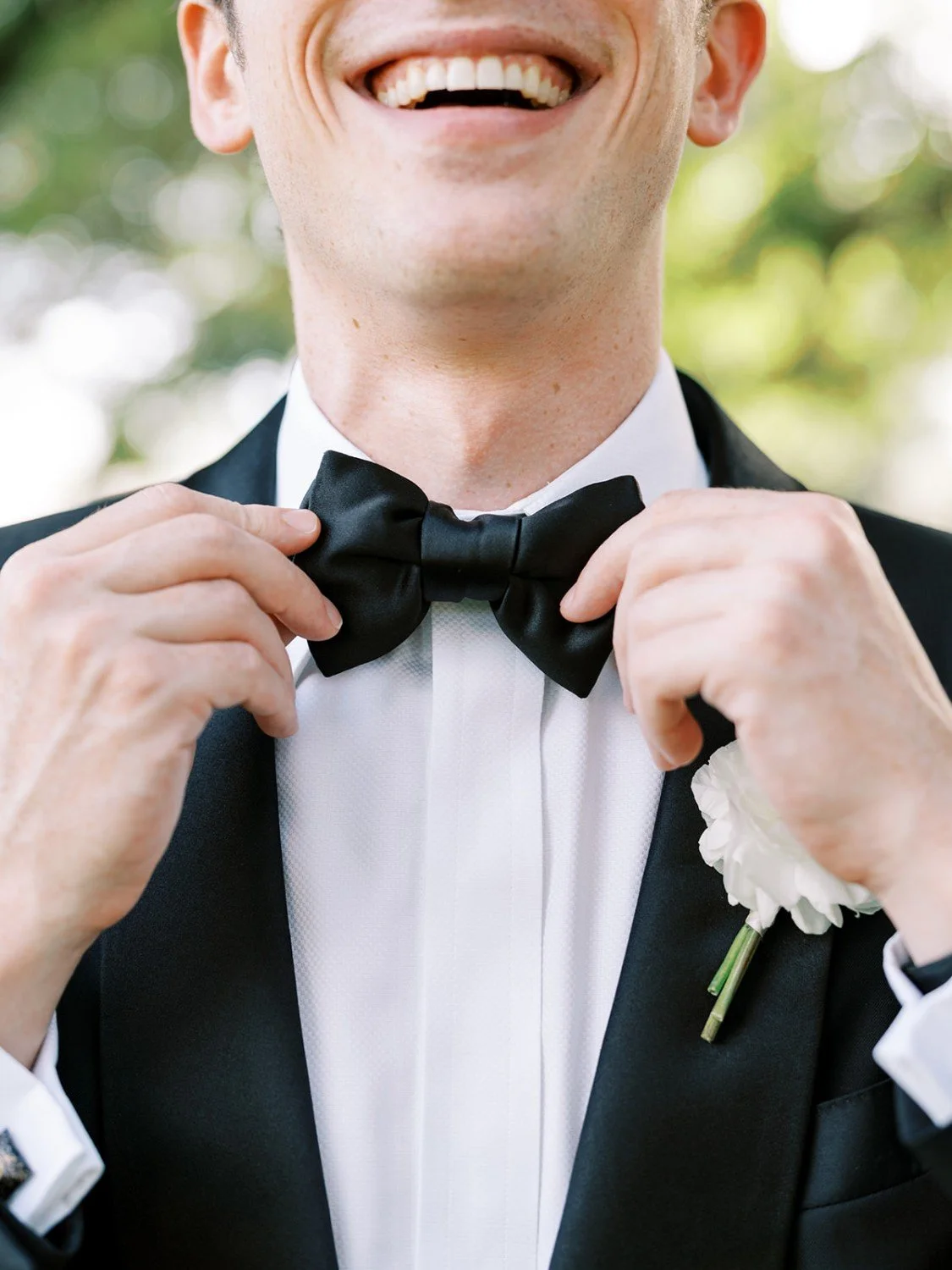 A man in a tuxedo adjusting a black bow tie with a white dress shirt and a white carnation boutonniere while smiling outdoors.