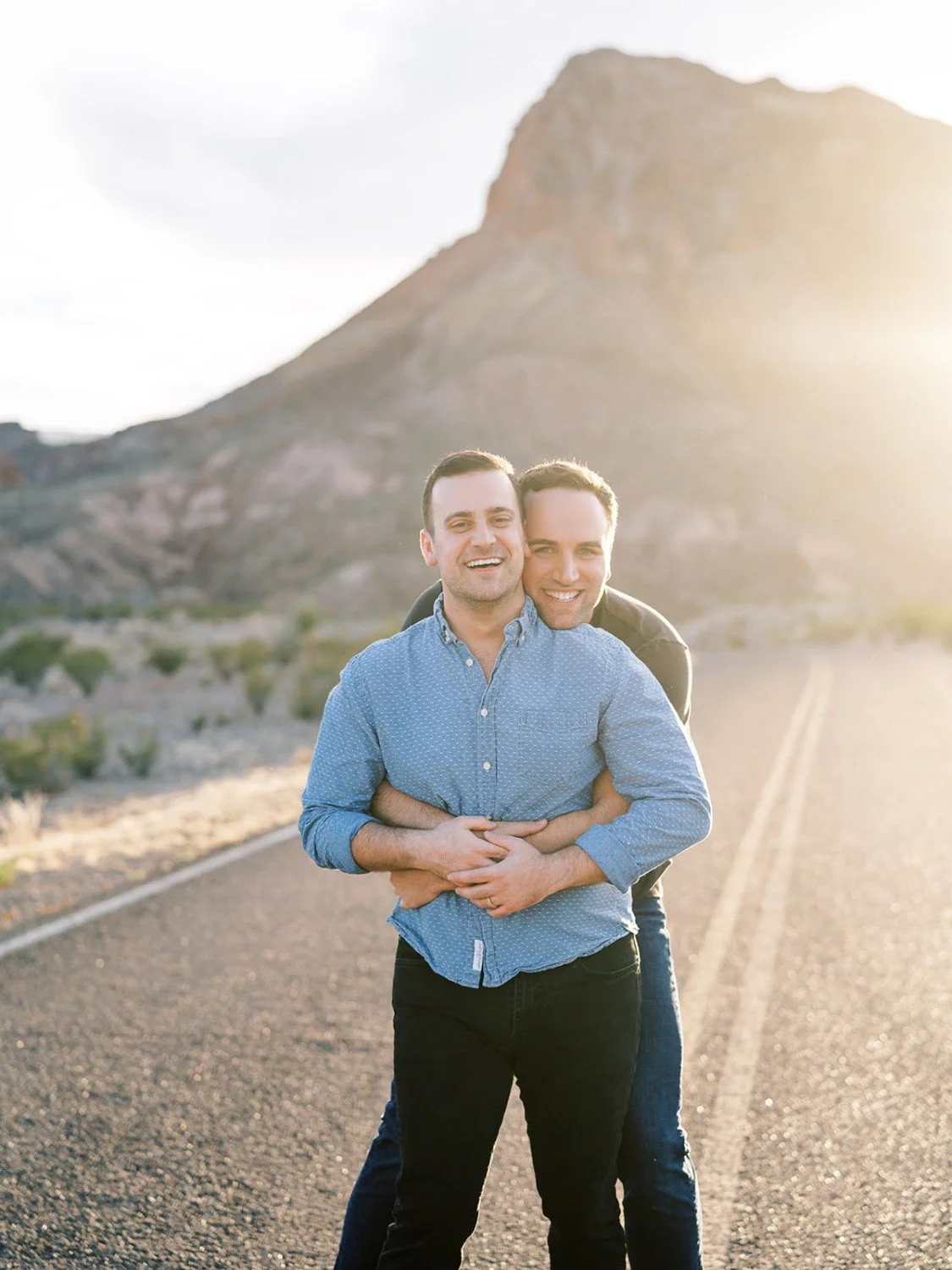 Two men happily hugging on an empty road with a mountain in the background at sunset