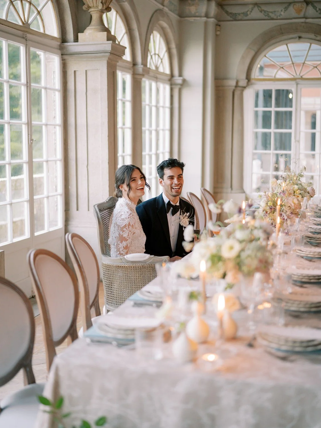 A bride and groom sitting at a decorated banquet table during a wedding celebration, smiling and enjoying the moment in a bright, elegant room with large windows and floral arrangements.