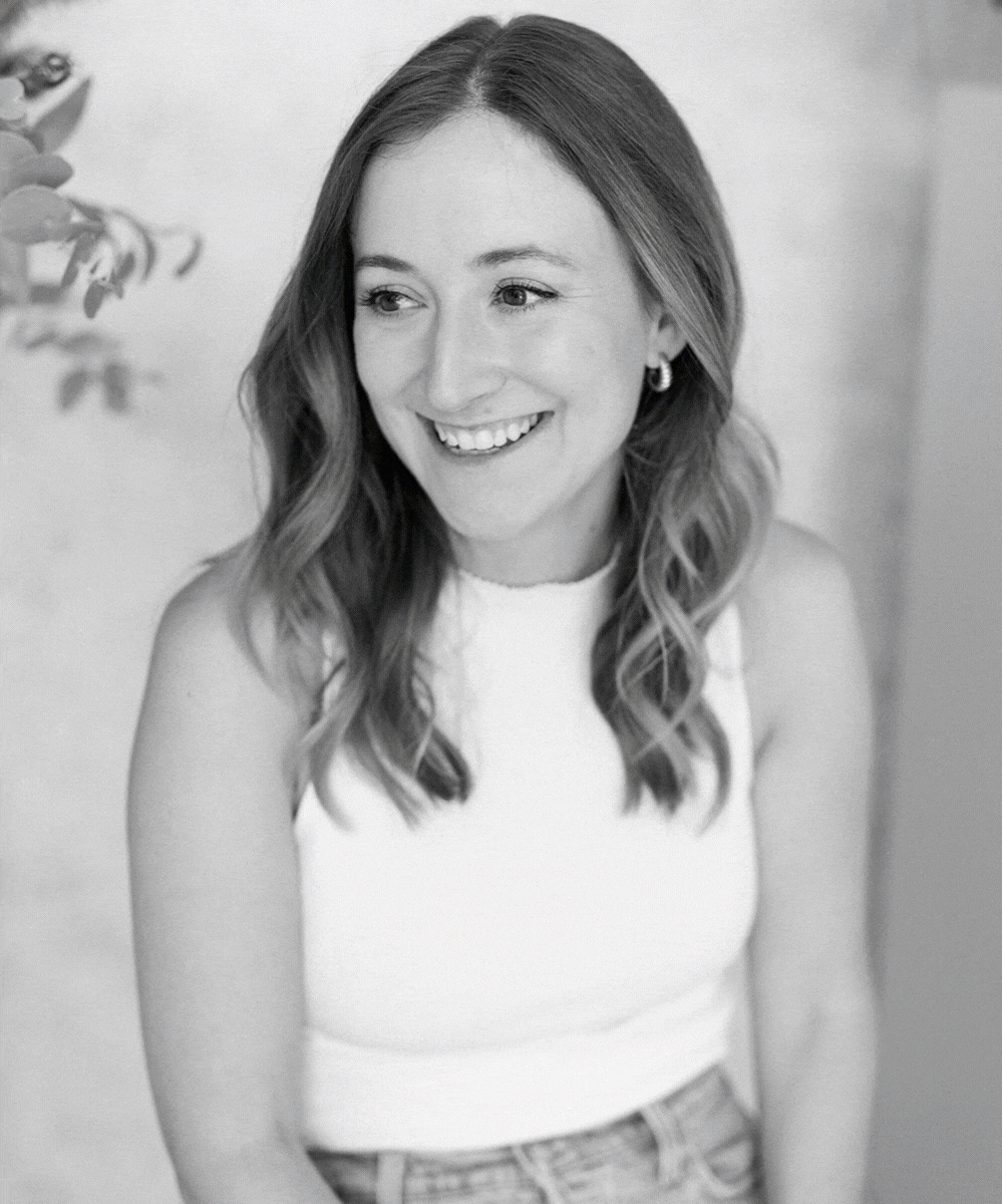 A smiling woman with wavy hair wearing a sleeveless top and earrings, standing indoors against a light background.