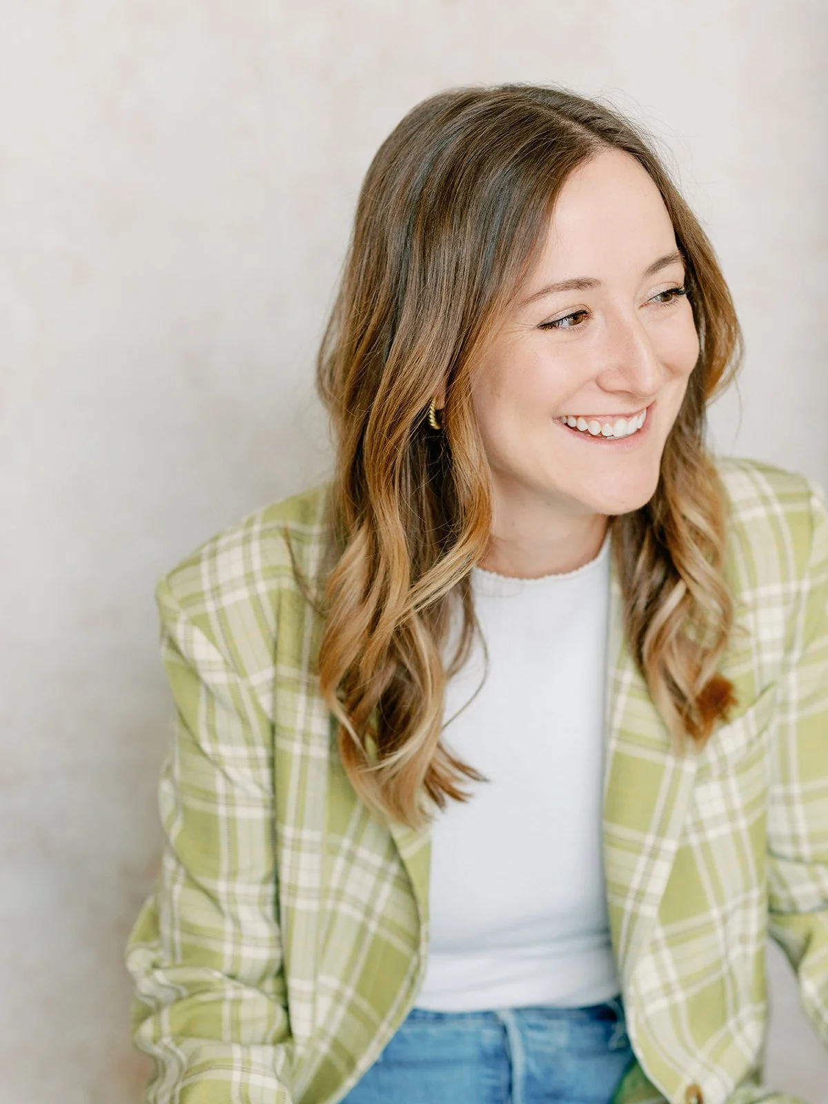 A young woman with light brown, wavy hair smiling and looking to her right, wearing a white top and a green checked blazer.