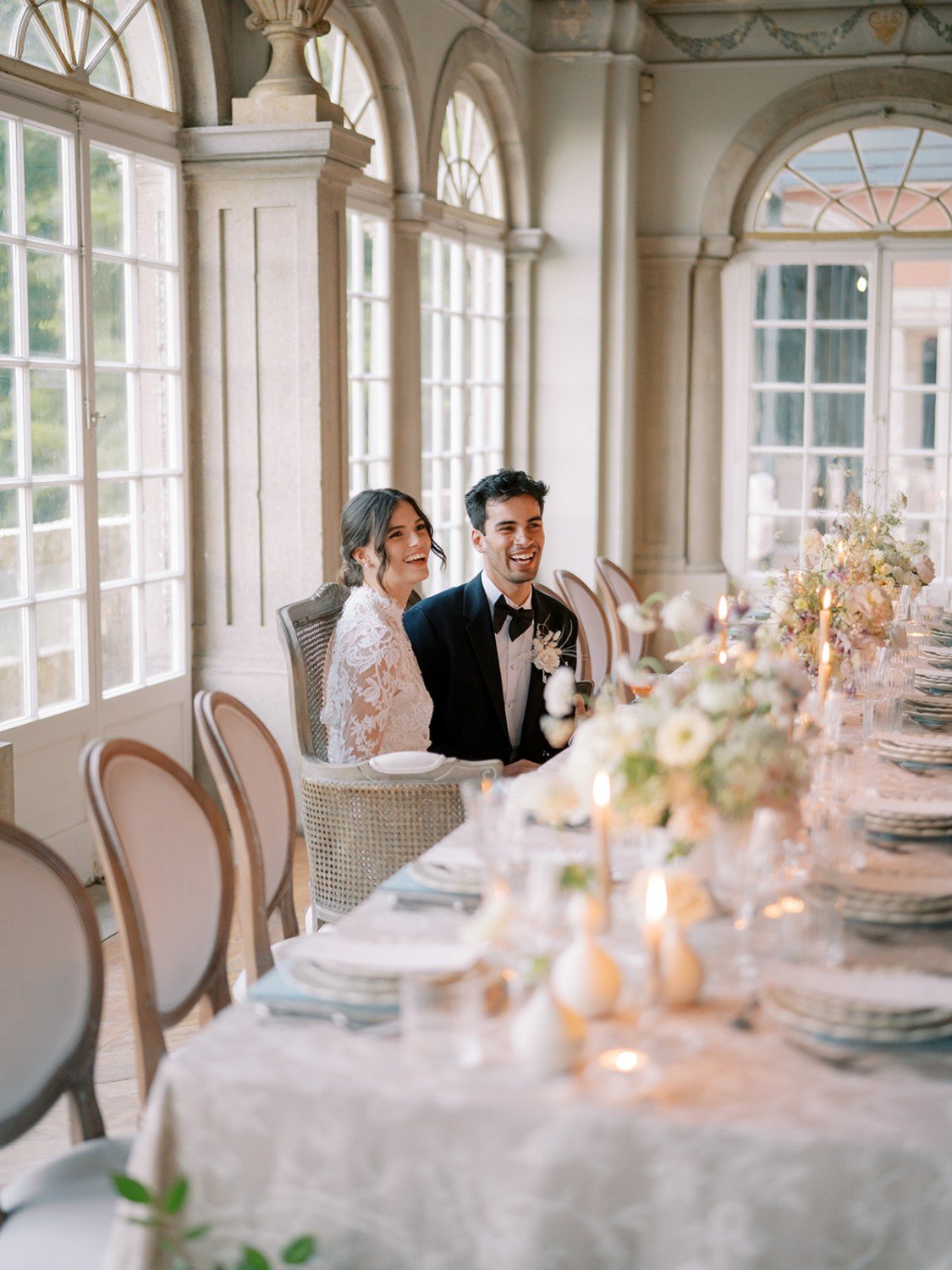 A bride and groom sitting at a decorated wedding reception table, smiling and laughing inside a bright, elegant room with large windows and floral arrangements.