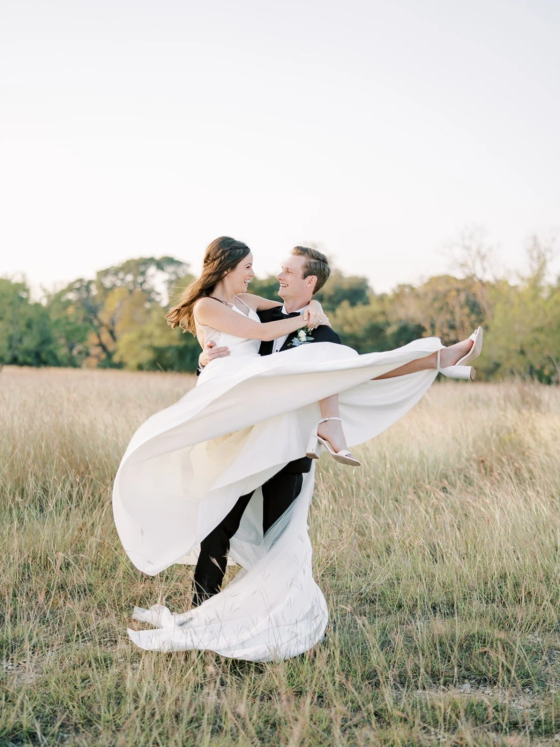 A bride and groom in wedding attire in a grassy field, with the groom lifting the bride in his arms, both smiling at each other.